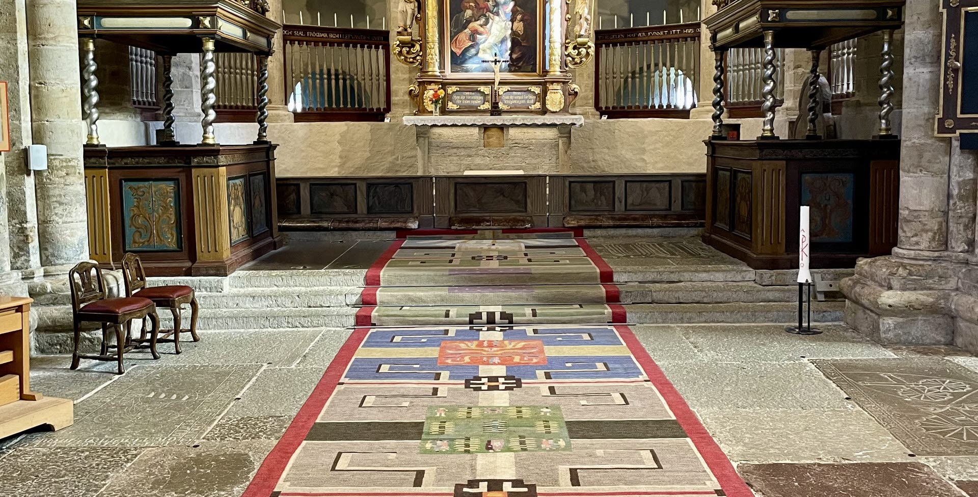 En vävd kormatta i ånga olika färger. Mattan ligger i en kyrka. På mattan står det Agda Österberg 1951
