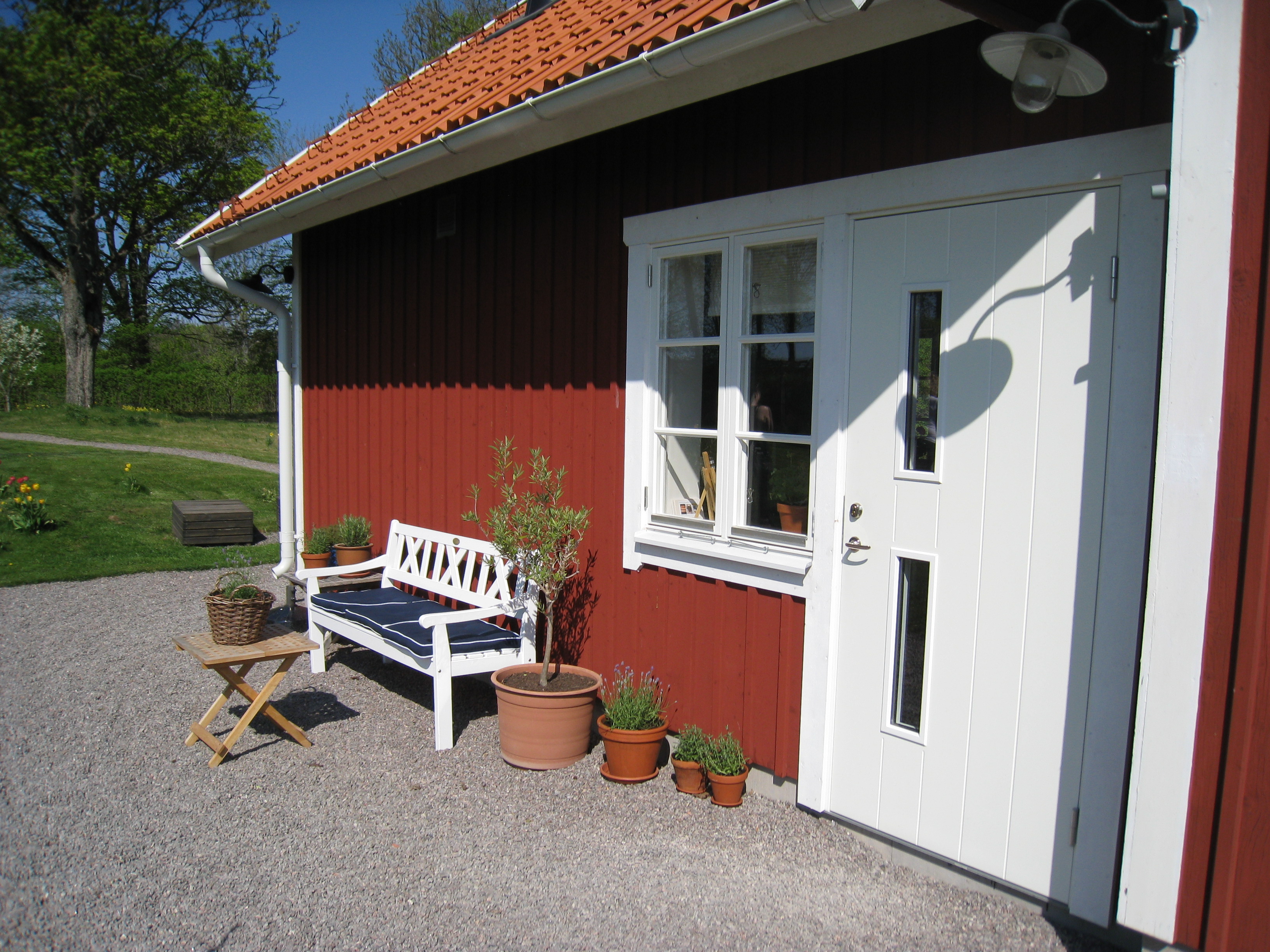 Ett rött hus med en vit dörr och ett fönster. Utanför står en träsoffa och några växter.