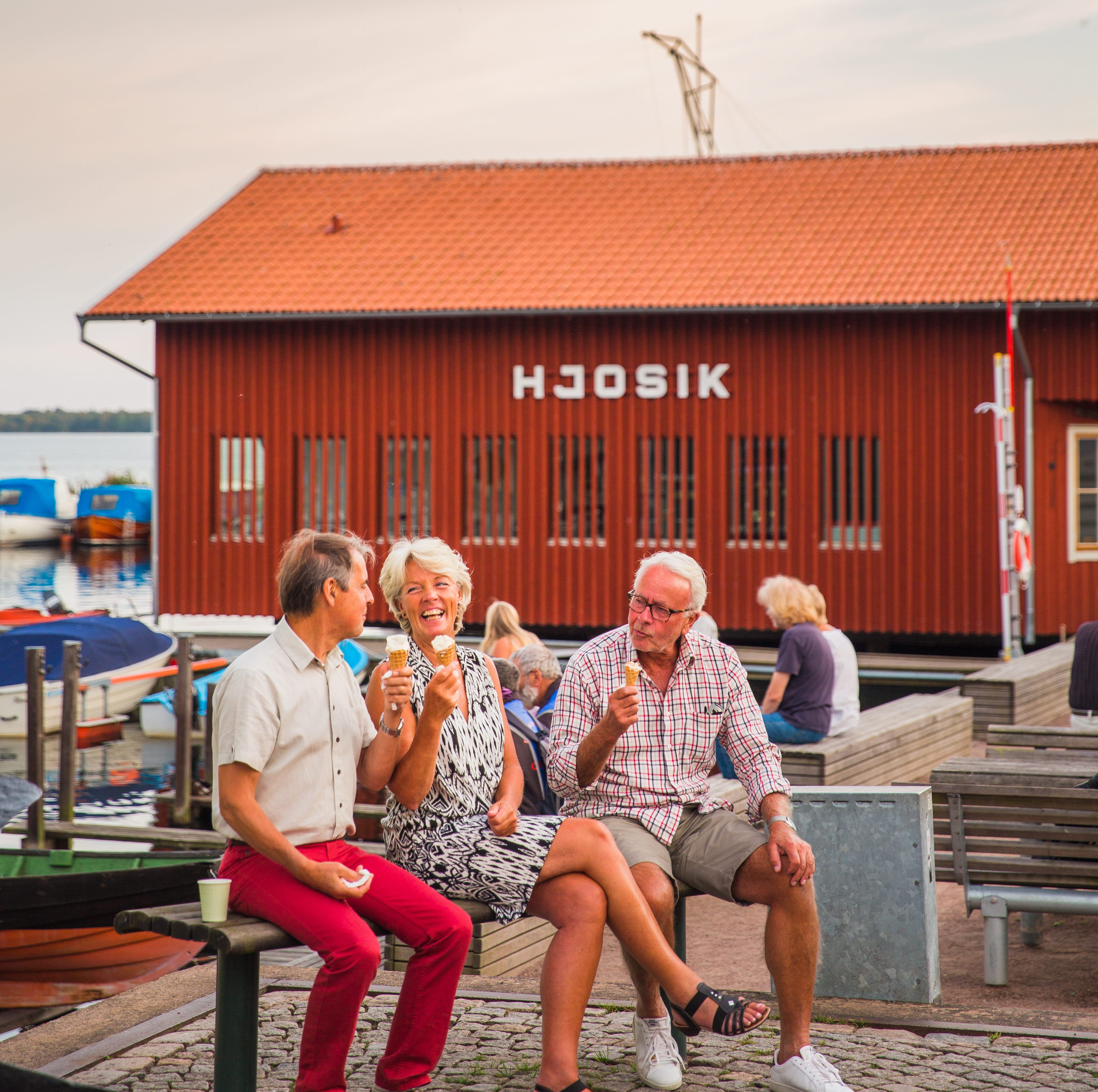 Tre människor äter glass i Hjo hamn, med fiskaffären Hjo Sik i bakgrunden. 