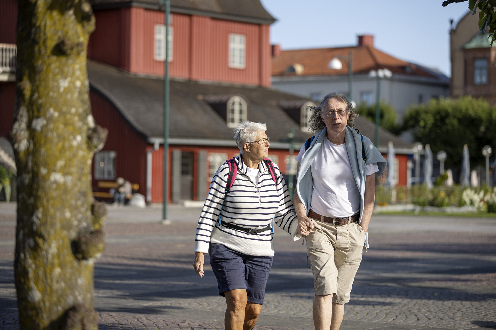 Ett äldre par håller varandra i handen på ett torg med ett rött hus i bakgrunden.
