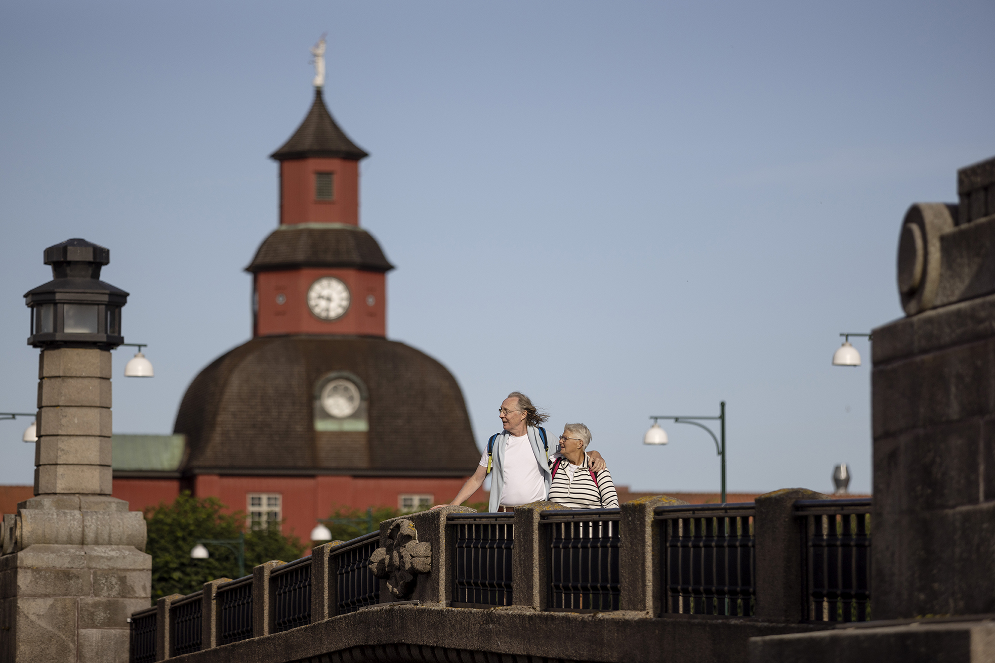 Ett äldre par står på en stenbro med en röd byggnad i bakgrunden.