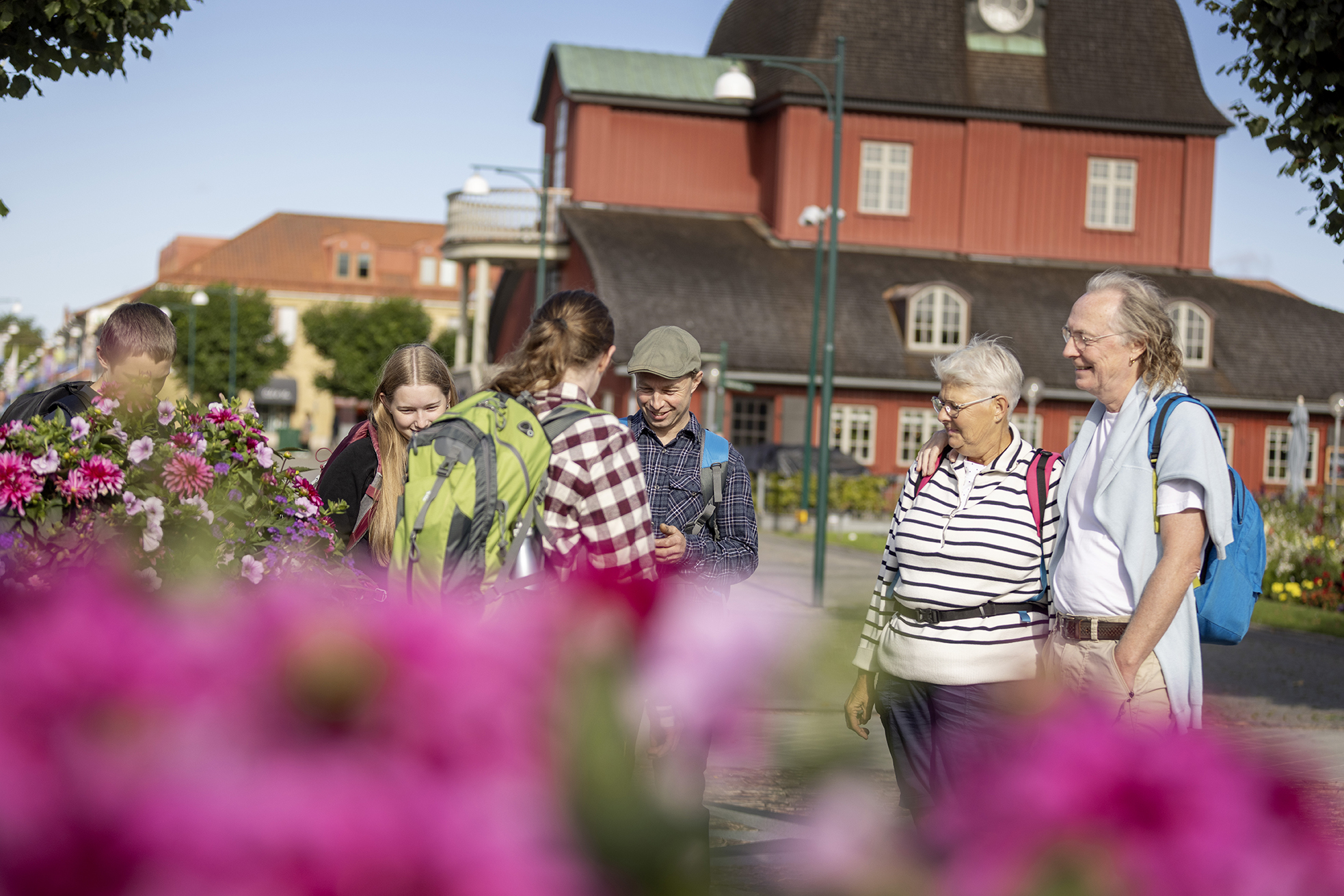 Det äldre paret och familjen står och pratar på torget. En röd byggnad i bakgrunden.
