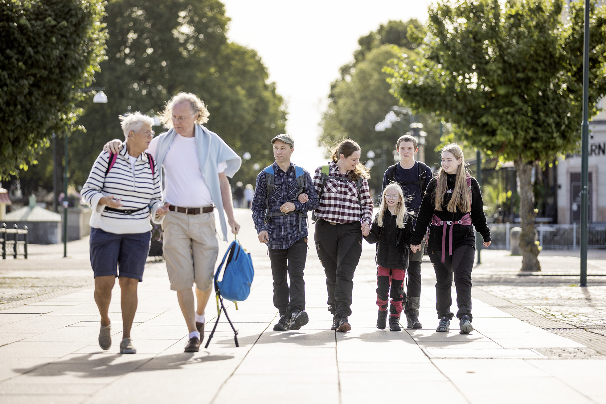 Ett äldre par och en familj med mamma, pappa och barn går på ett torg. Det finns gröna träd i bakgrunden.