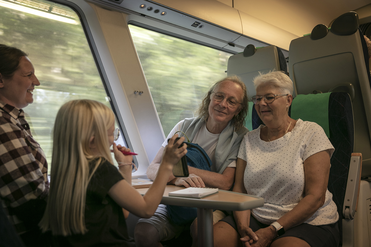 De äldre paret sitter mitt emot en mamma och hennes flicka som visar upp något på sin mobiltelefon.