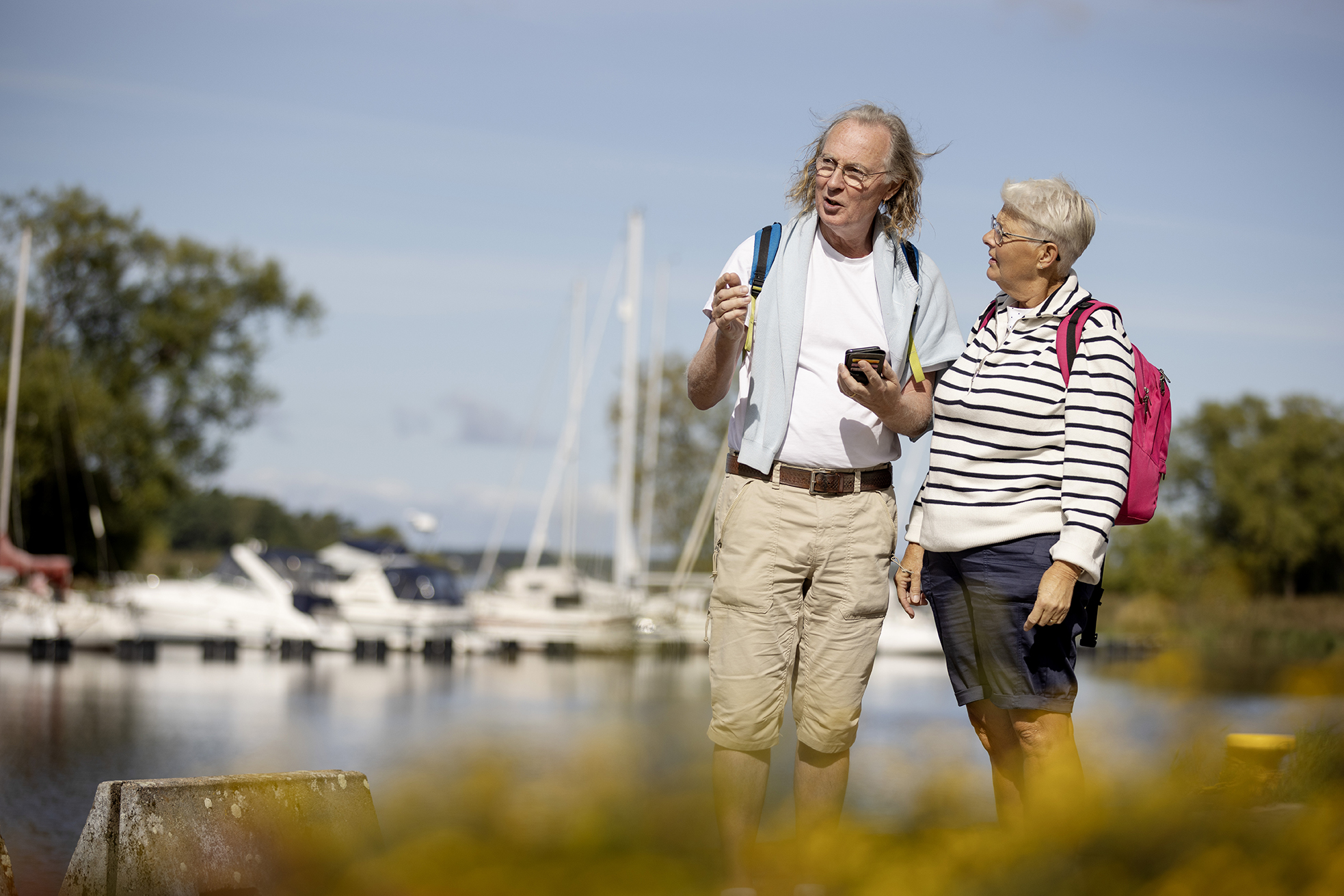 Ett äldre par tittar åt vänster i bilden. Det finns en marina med båtar i bakgrunden.