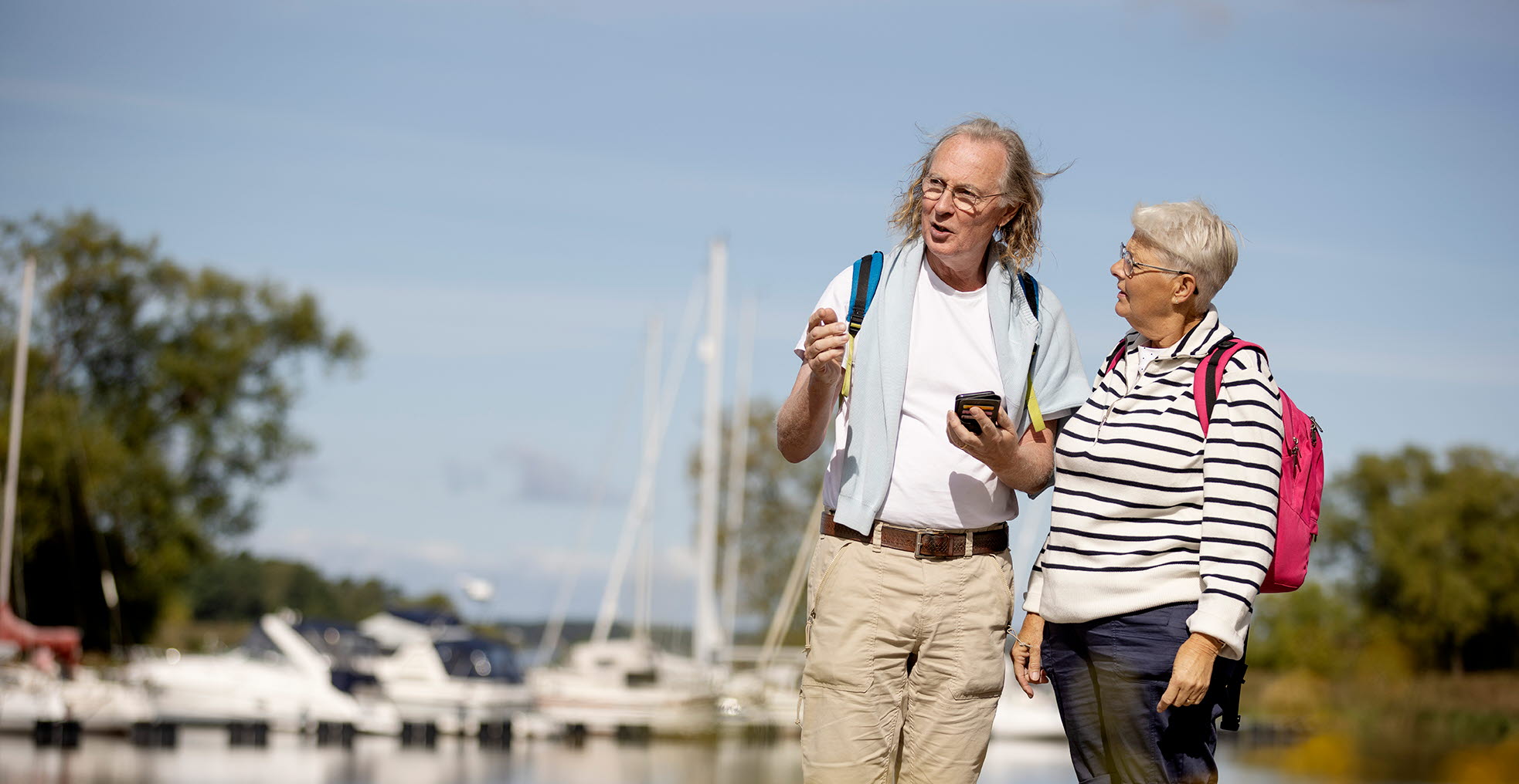 Ett äldre par tittar åt vänster i bilden. Det finns en marina med båtar i bakgrunden.
