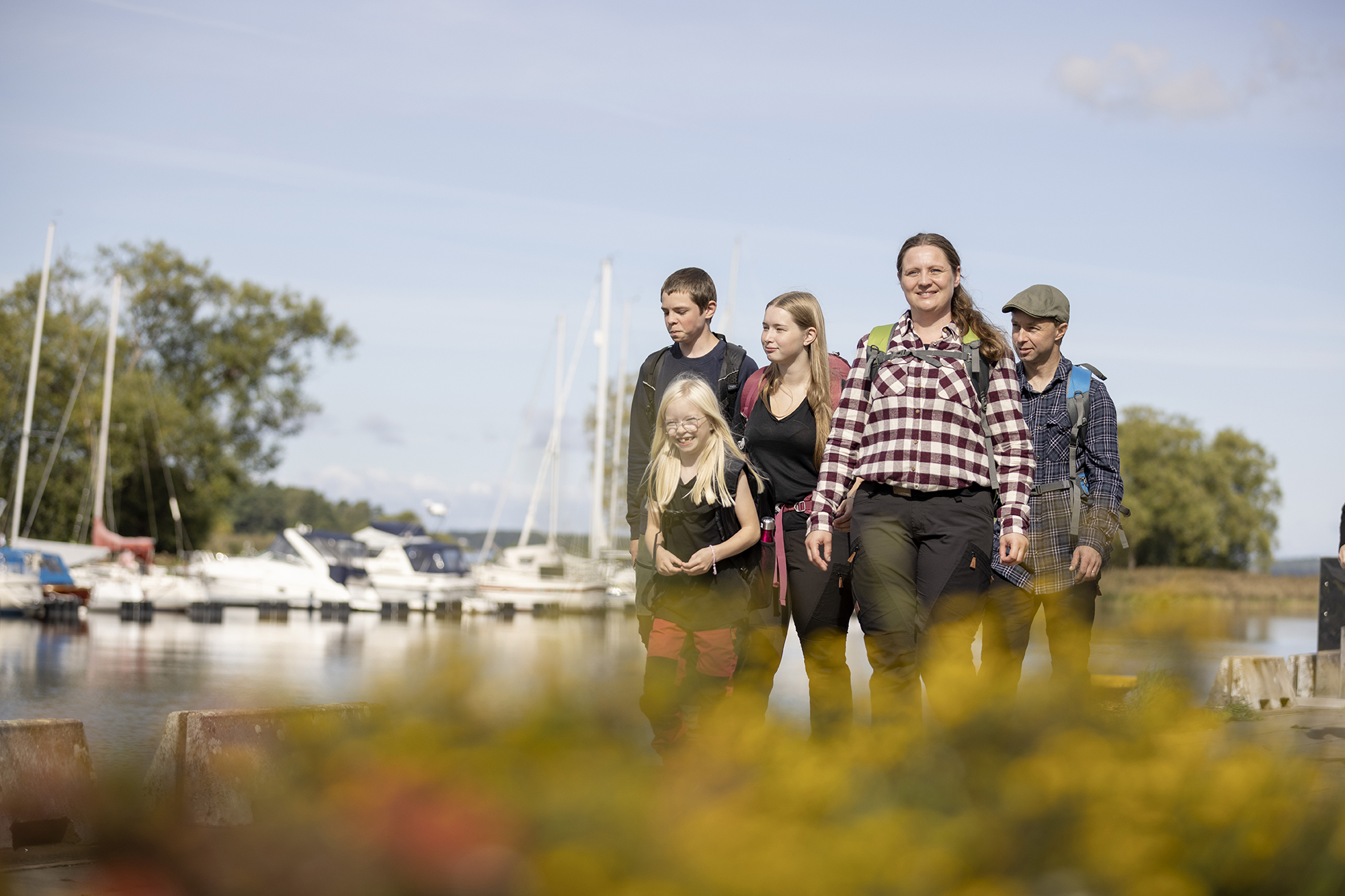 Familj med mamma, pappa och tre barn står och tittar åt vänster i bilden. Det är en båthamn med båtar i bakgrunden.