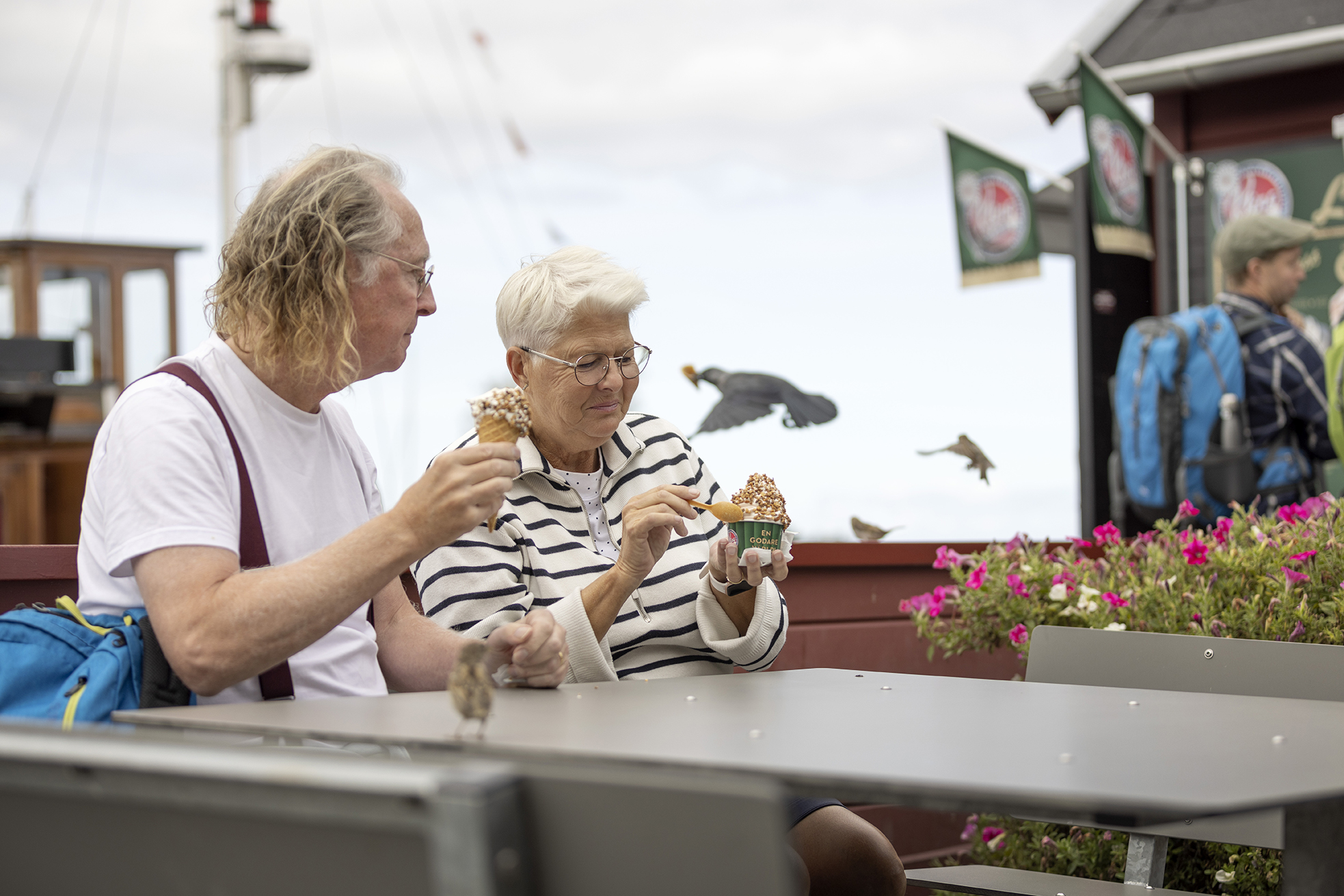 Ett äldre par sitter ner vid ett bord och äter glass. Det finns en gammal träbåt i bakgrunden.