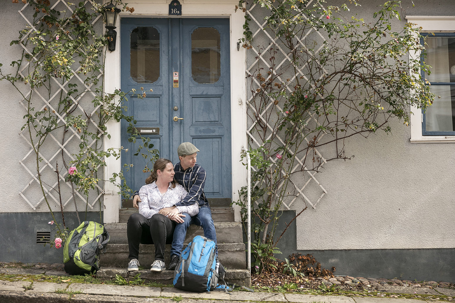 En man och en kvinna sitter och håller om varandra på en trappa till ett vitt hus med en blå dubbeldörr. Deras ryggsäckar står på marken framför trappan.