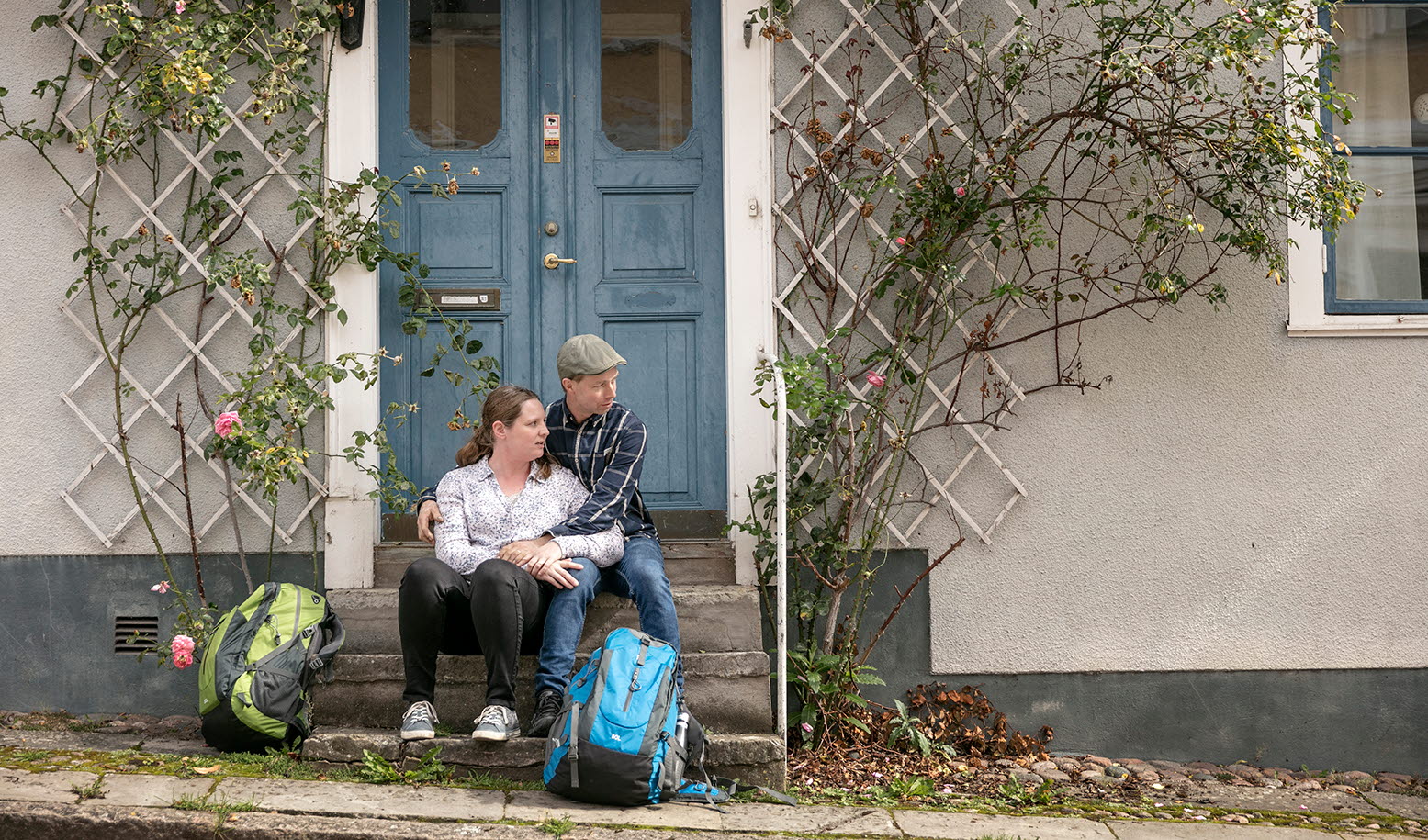 En man och en kvinna sitter och håller om varandra på en trappa till ett vitt hus med en blå dubbeldörr. Deras ryggsäckar står på marken framför trappan.