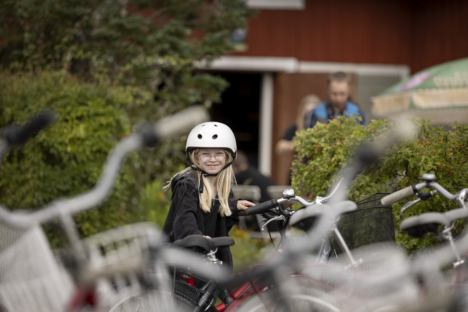 En glad blond tjej står och håller i en cykel.
Hon har en vit cykelhjälm.