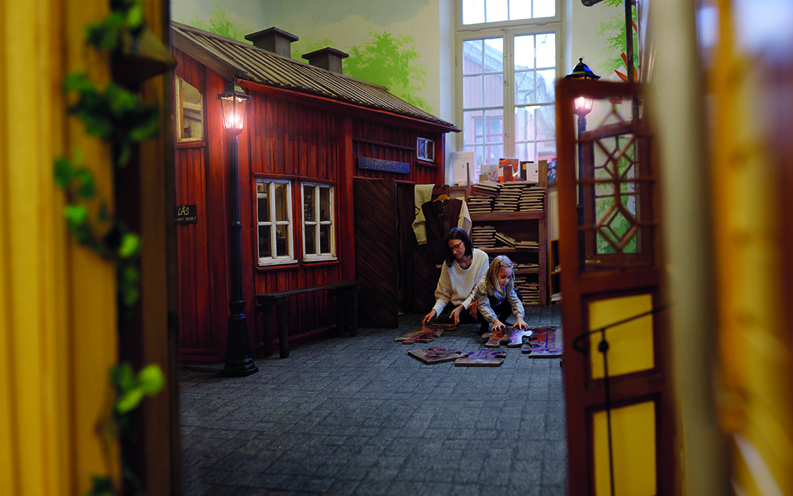 Vy inifrån Bokäventyret, en mamma och hennes barn leker på golvet framför en byggnad av miniformat