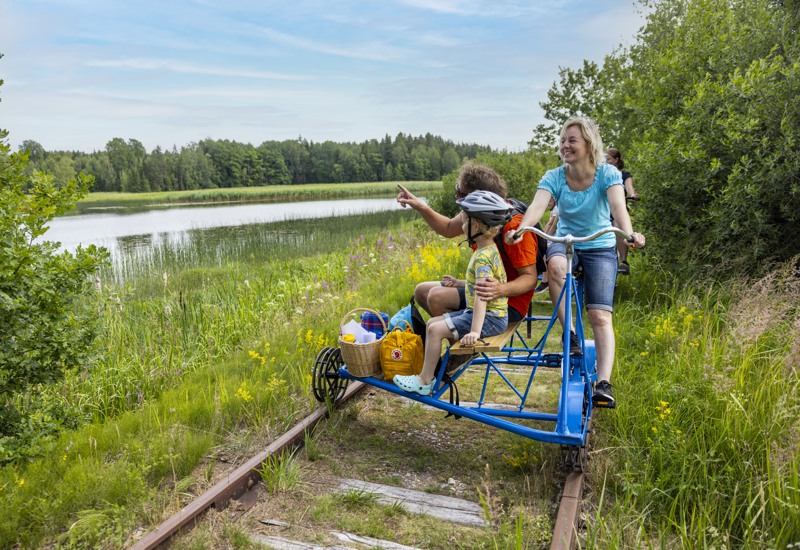 En kvinna i blå t-shirt, en man i röd t-shirt och ett barn med cykelhjälm sitter på en dressin och tittar ut över en sjö.
