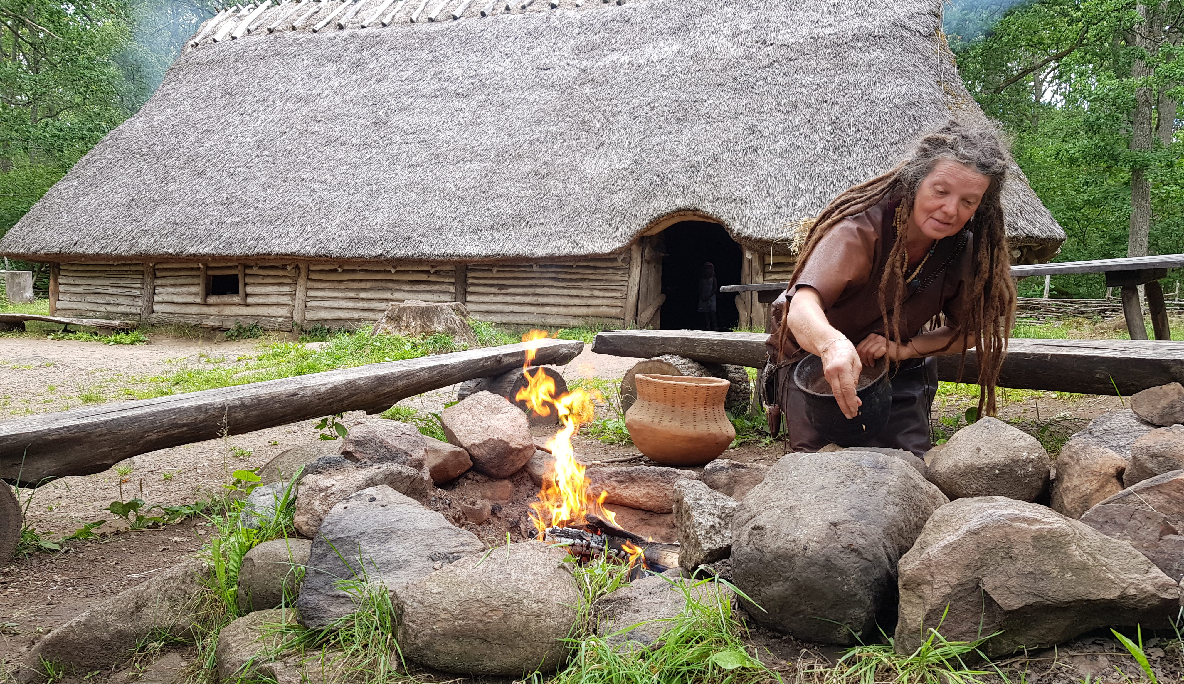Kvinna som rostar säd vid en öppen eld. I bakgrunden syns ett trähus med vasstak.