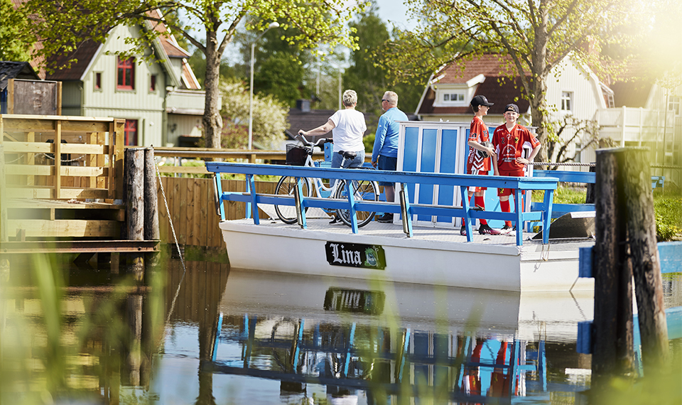 Sveriges minsta färja som hjälper boende och turister att korsa Göta kanal i Töreboda. På färjan syns en kvinna och man med cykel samt två unga killar i fotbollskläder.
