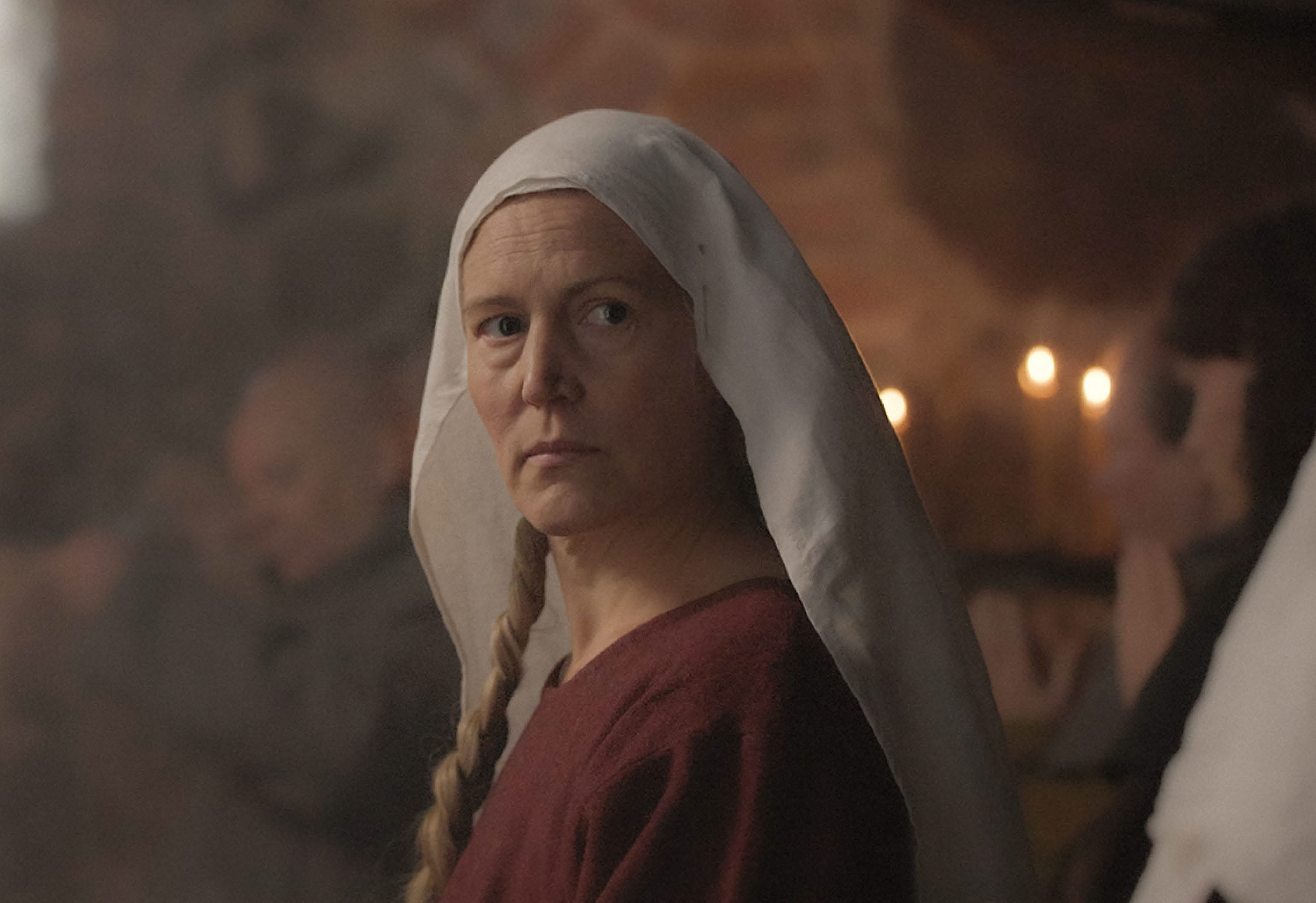 En kvinna i en gammeldags dräkt med dok. Hon står i en kyrkliknande byggnad med tända ljus.