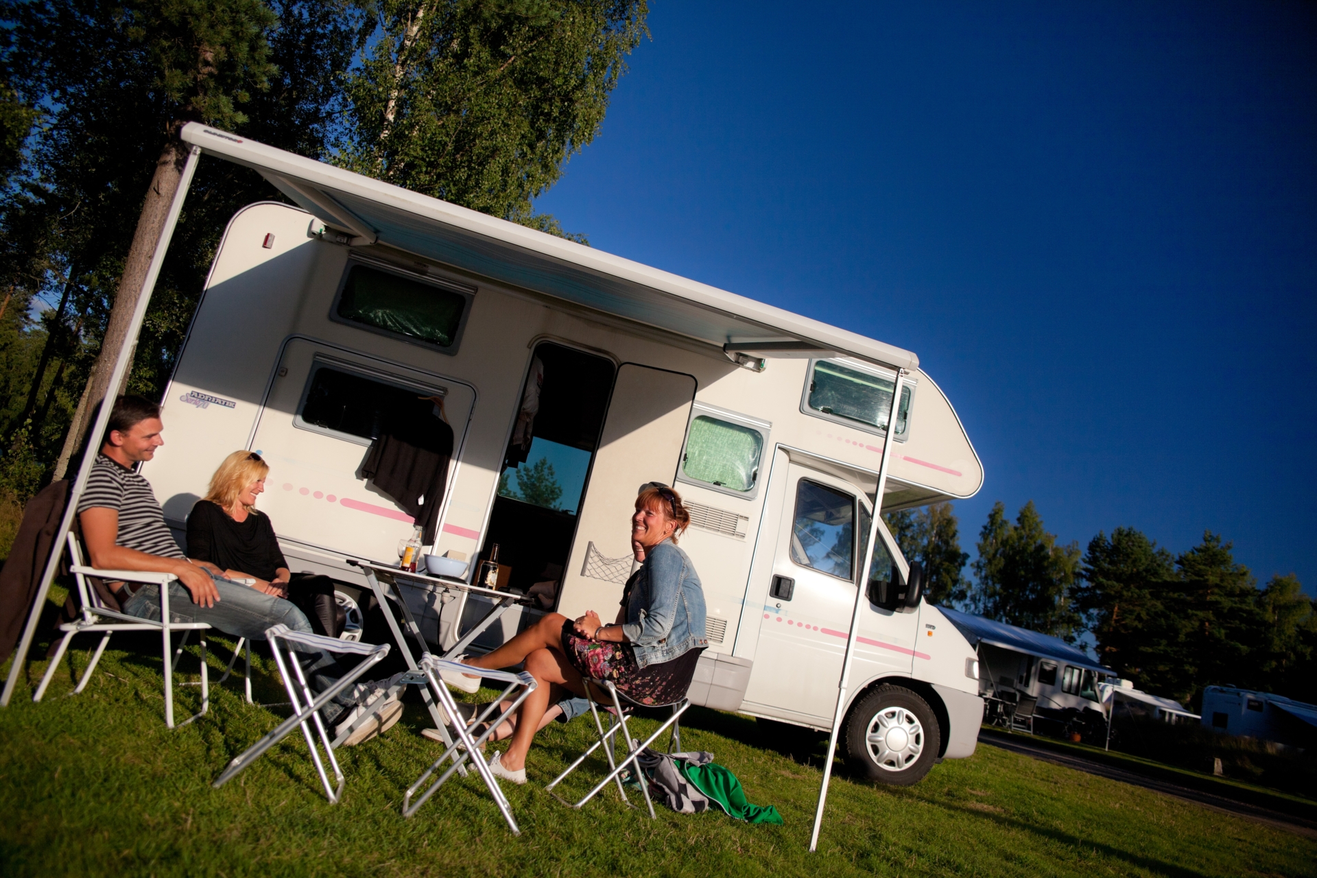 Husbil på Skara Sommarlands Camping. Tre personer sitter utanför.