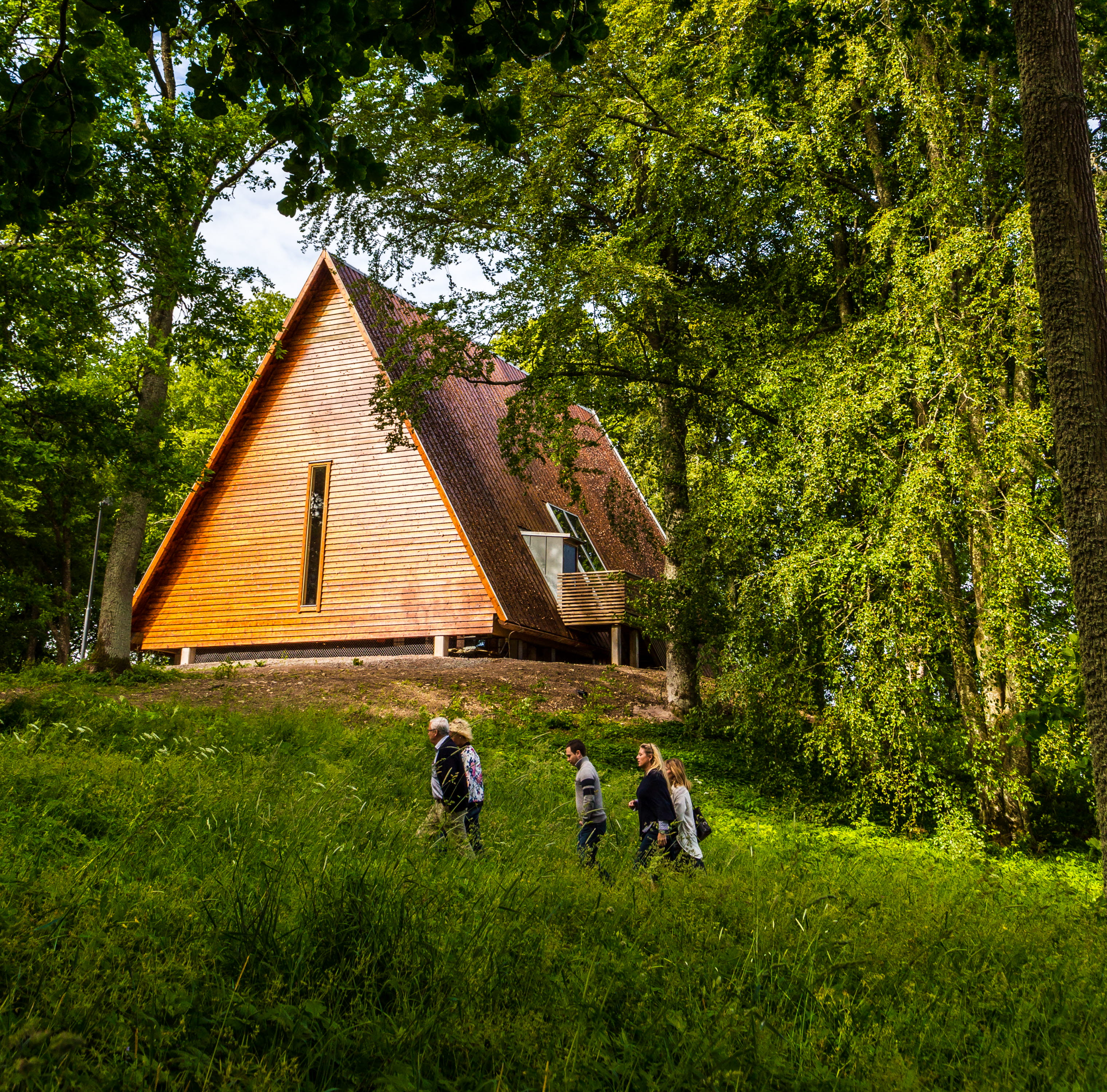 Kata gård, ett trähus på en kulle men grönskande träd omkring. Tre människor vandrar upp mot huset.