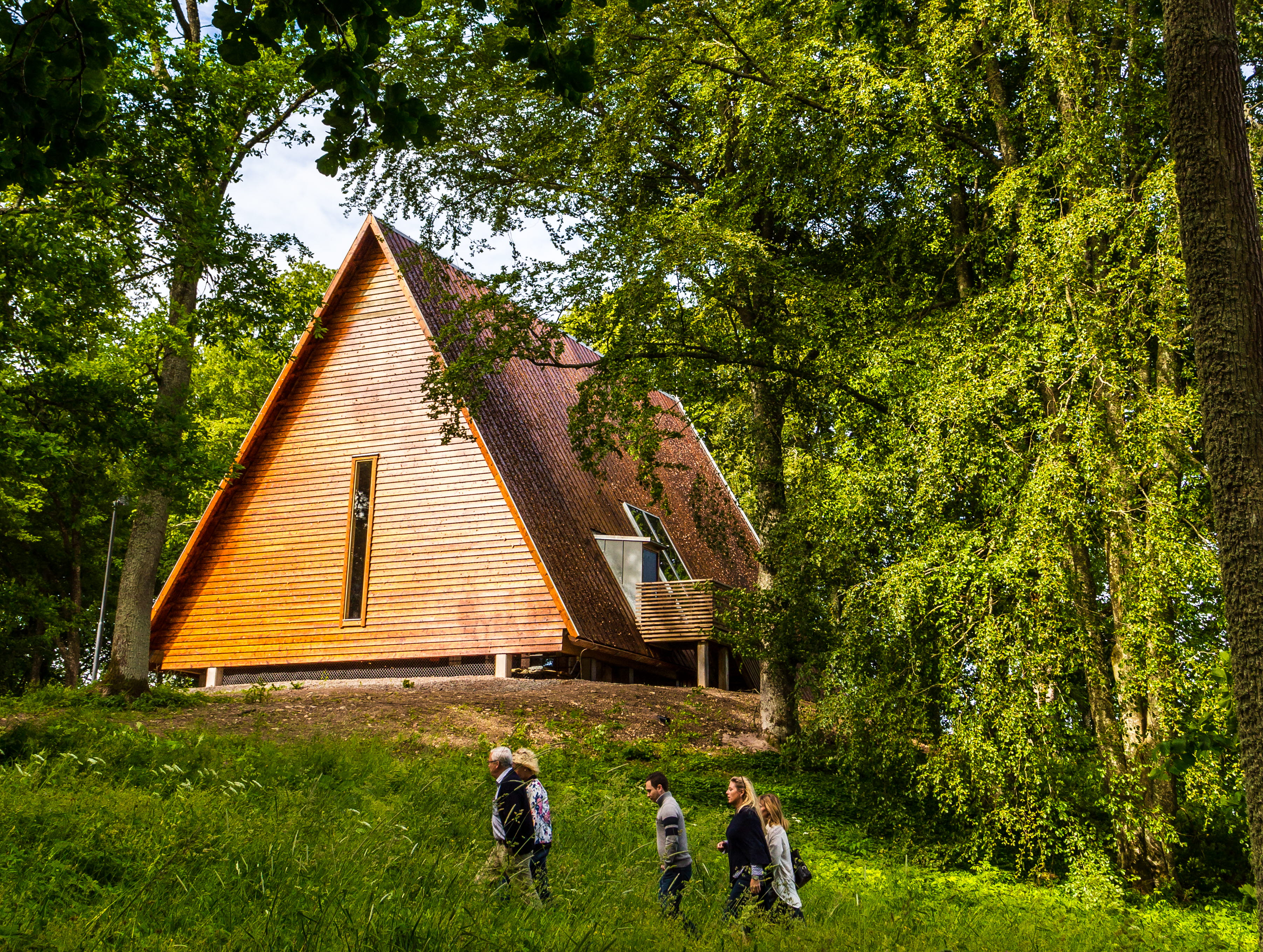 Kata gård, ett trähus på en kulle men grönskande träd omkring. Tre människor vandrar upp mot huset.