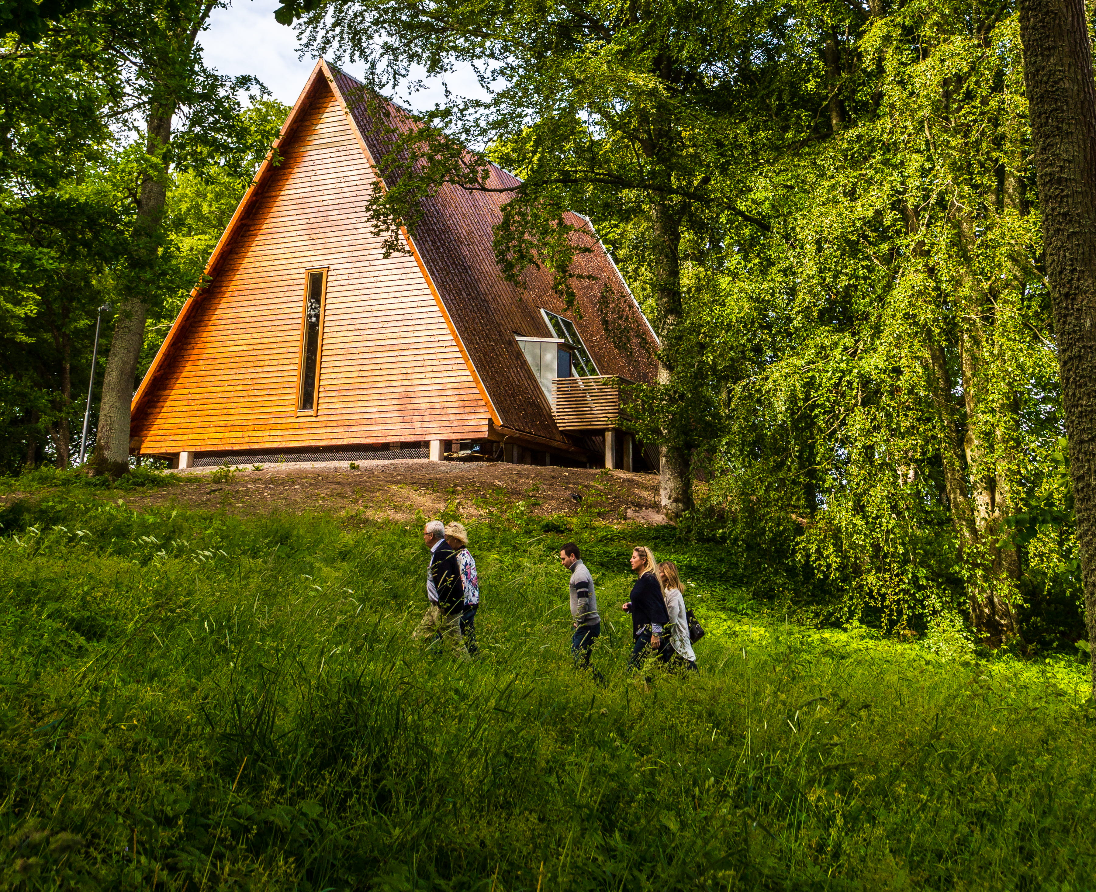 Kata gård, ett trähus på en kulle men grönskande träd omkring. Tre människor vandrar upp mot huset.