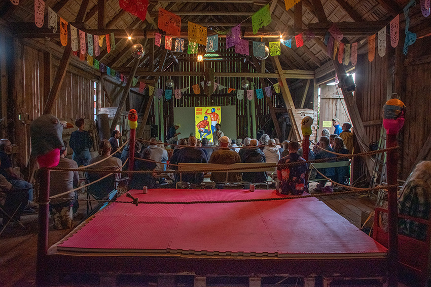 En lada som är omgjord till en sal för föreläsningar och uppvisningar. I Bakgrunden syns en scen med en projektorduk med ett bildspel. Bakom publiken syns en boxningsring med rött golv. I Taket hänger vimplar i olika färger.