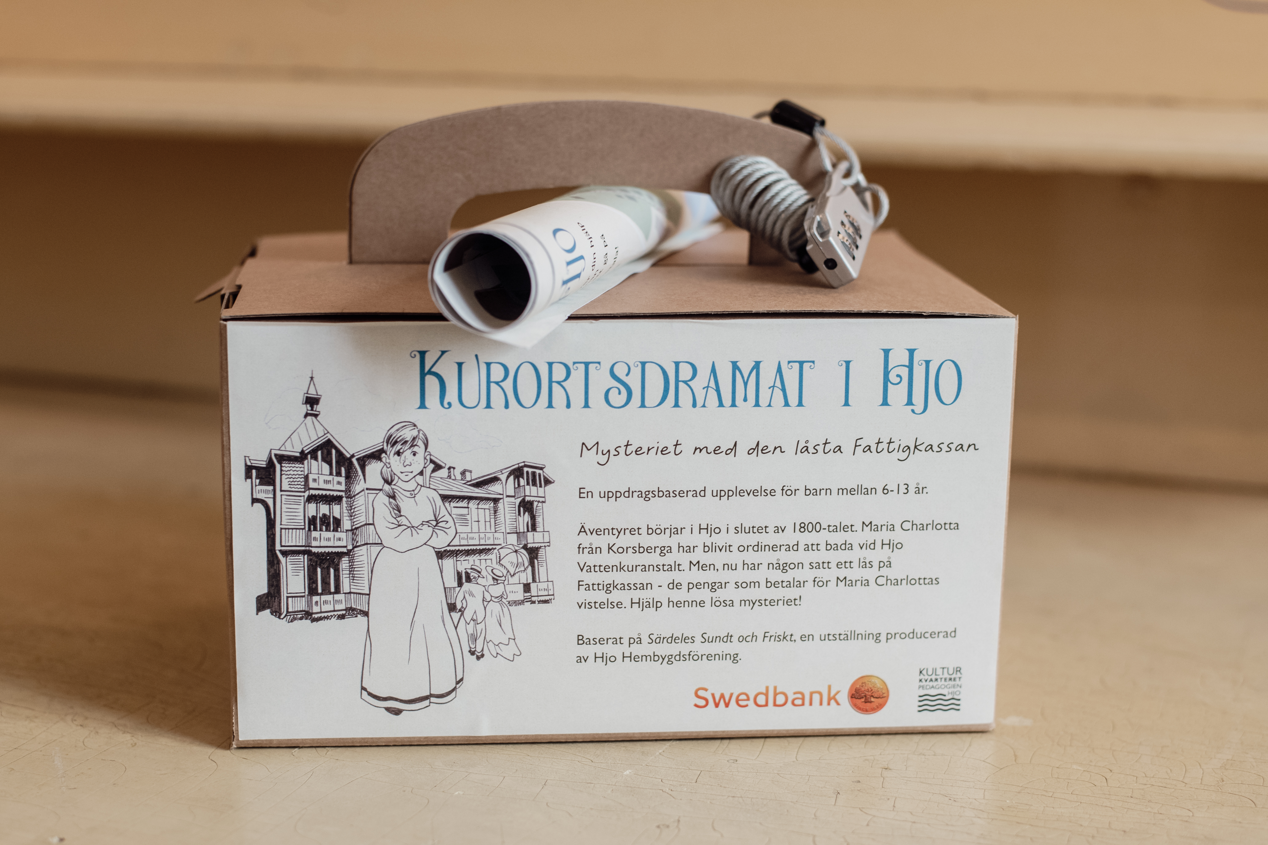 En liten låda med en pappersrulle ovanpå. På lådan finns en tecknad bild på en ung kvinna i en 1800-talsmiljö och en text där det står "Kurortsdramat" på. 