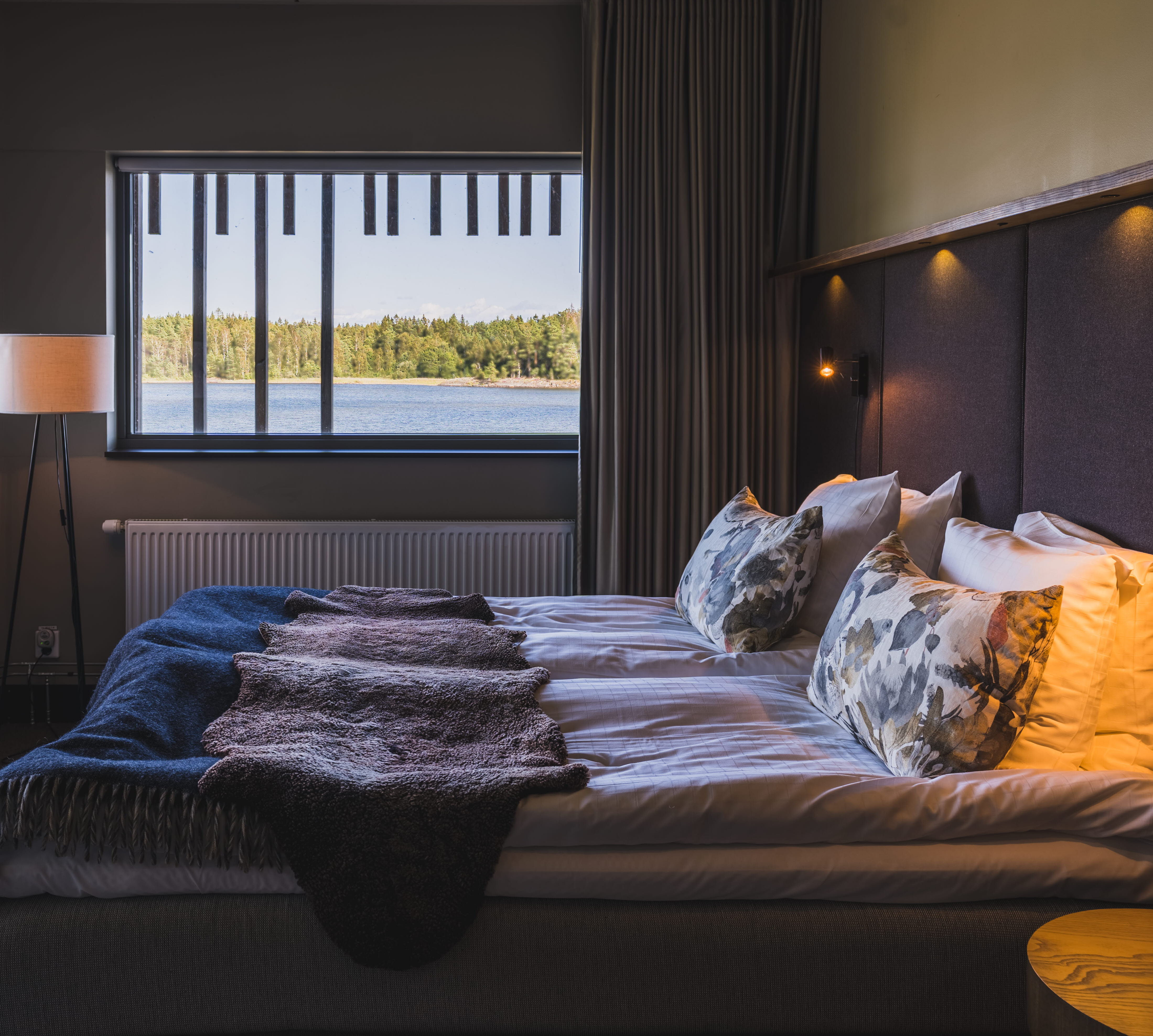 A hotel room with a double bed and a view of a lake through two windows.
