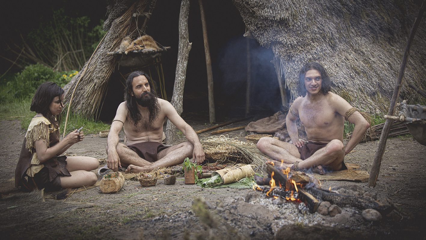 En flicka och två män sitter framför en hydda vid en lägereld