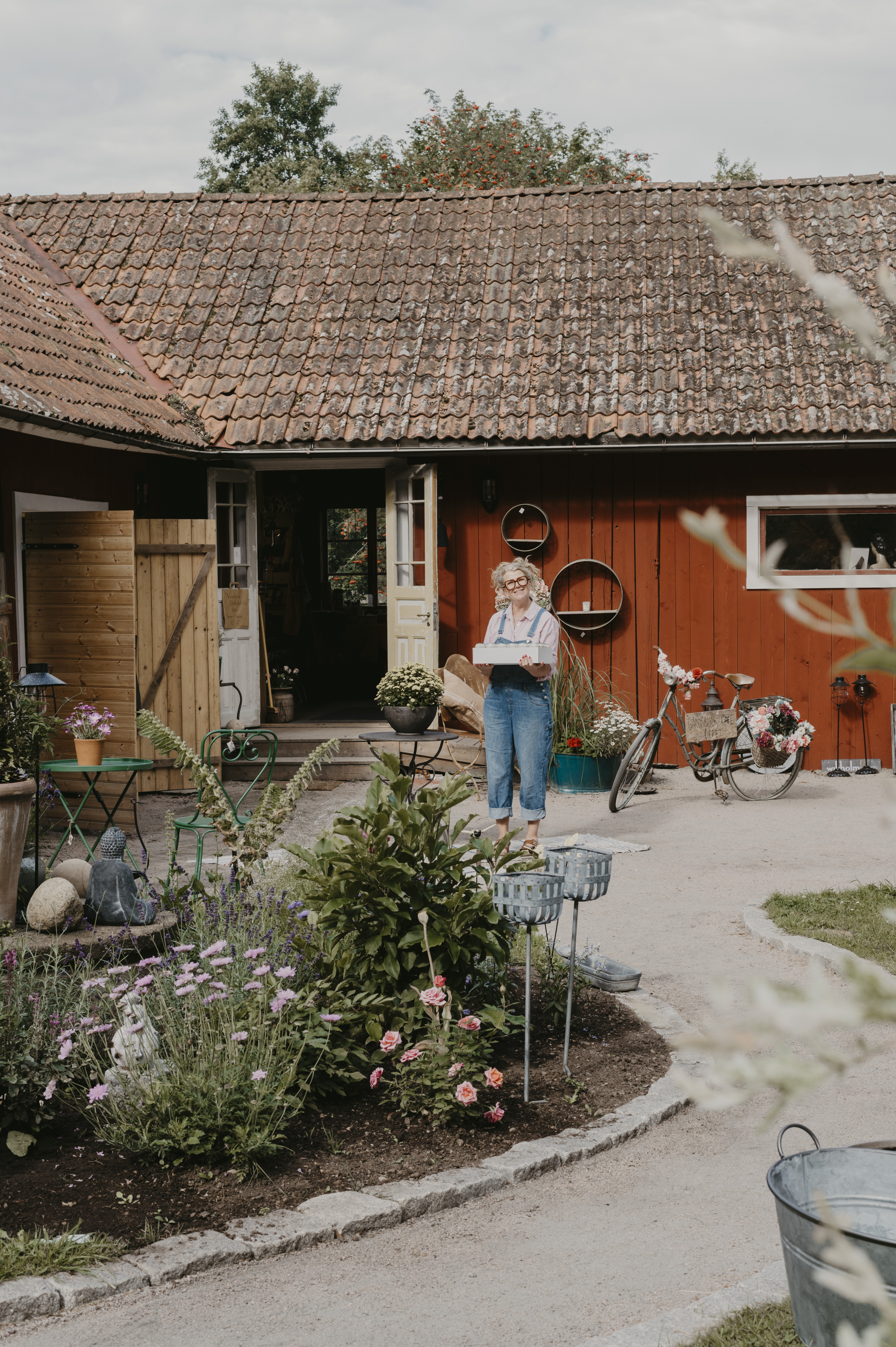 En kvinna håller i en låda. Hon har blå hängselbyxor på sig och står på en innergård. I förgrunden ser man en fin plantering.