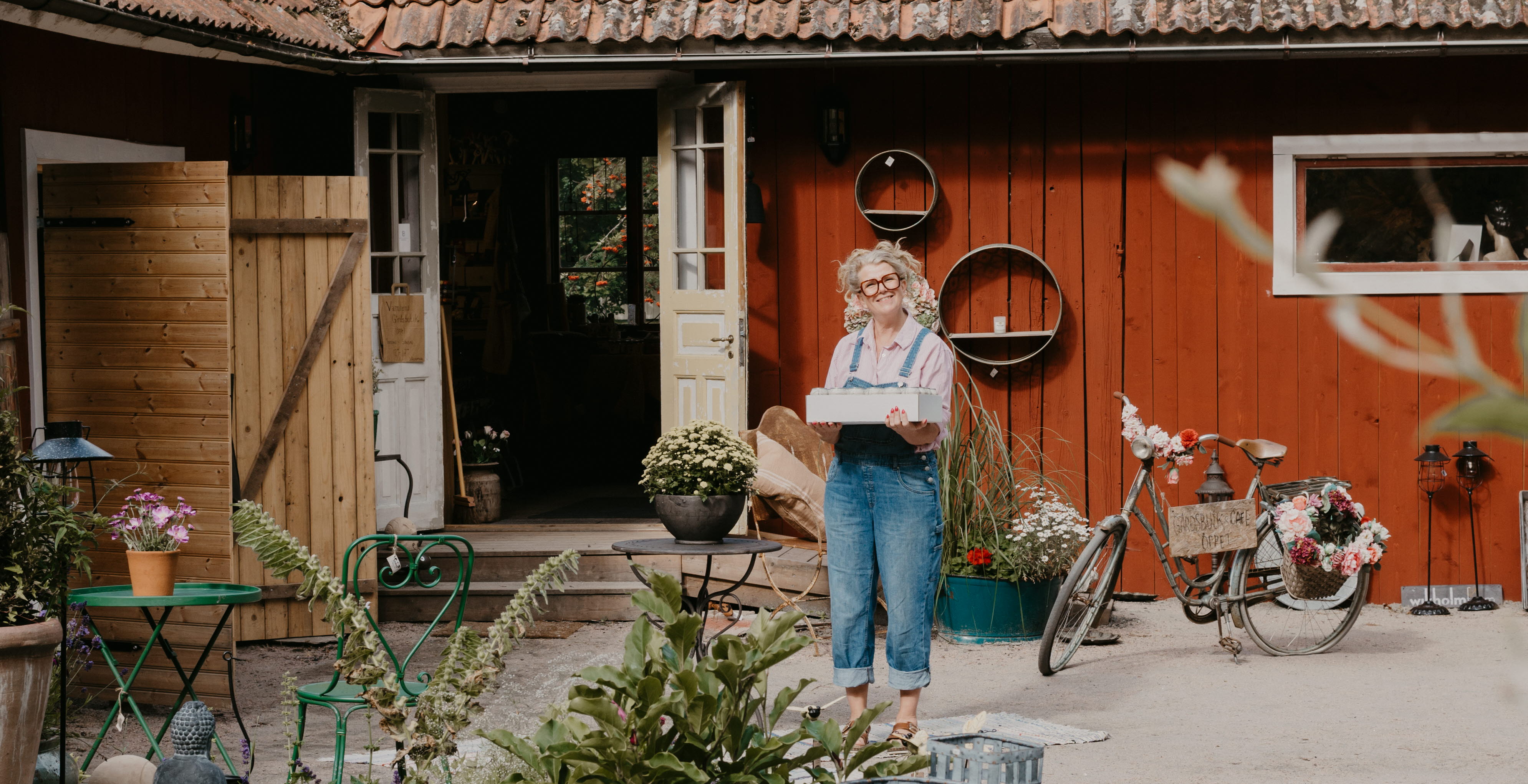 A woman is holding a box. She is wearing blue suspenders and is standing in a courtyard. In the foreground you can see a nice planting.
