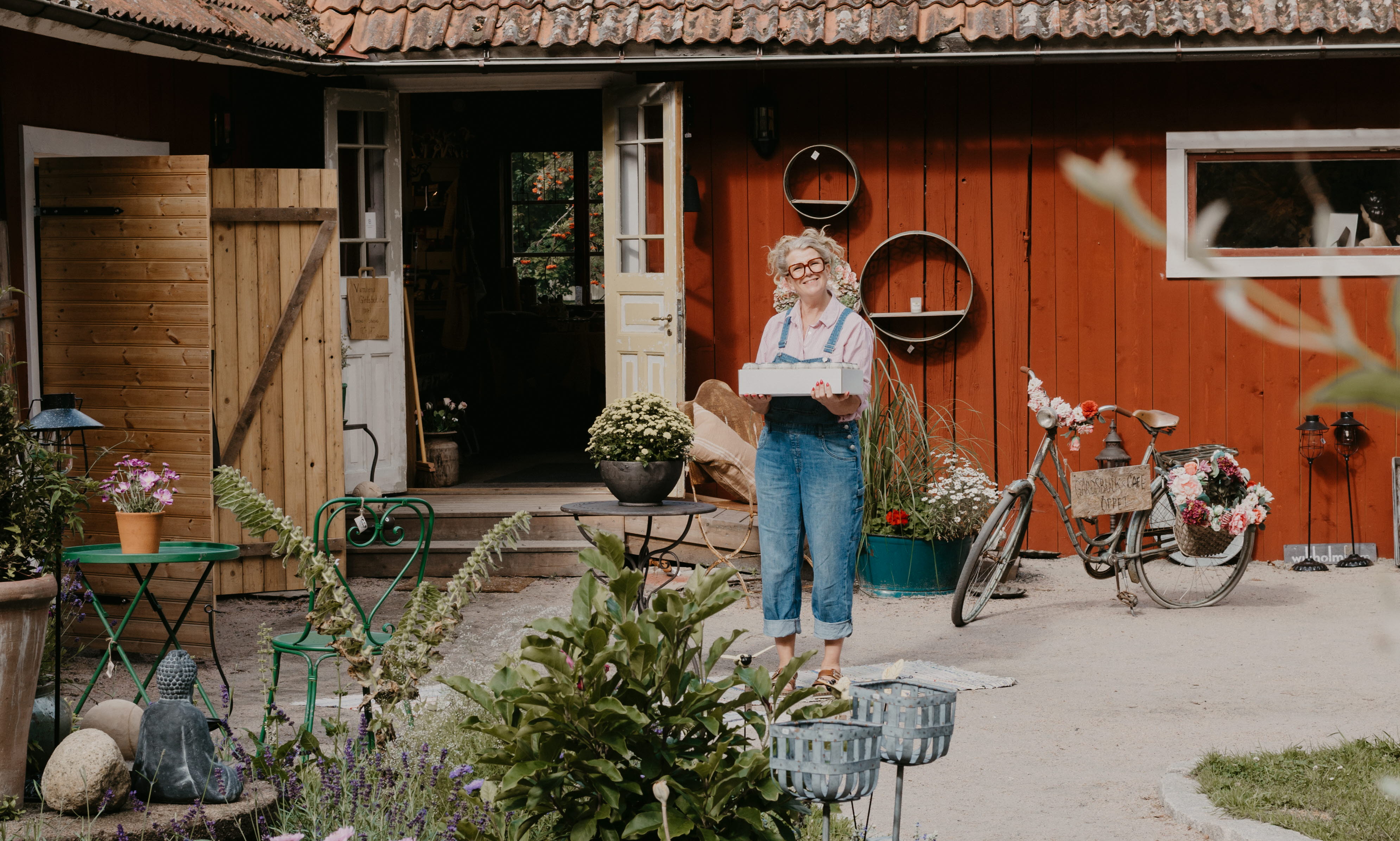 En kvinna håller i en låda. Hon har blå hängselbyxor på sig och står på en innergård. I förgrunden ser man en fin plantering.