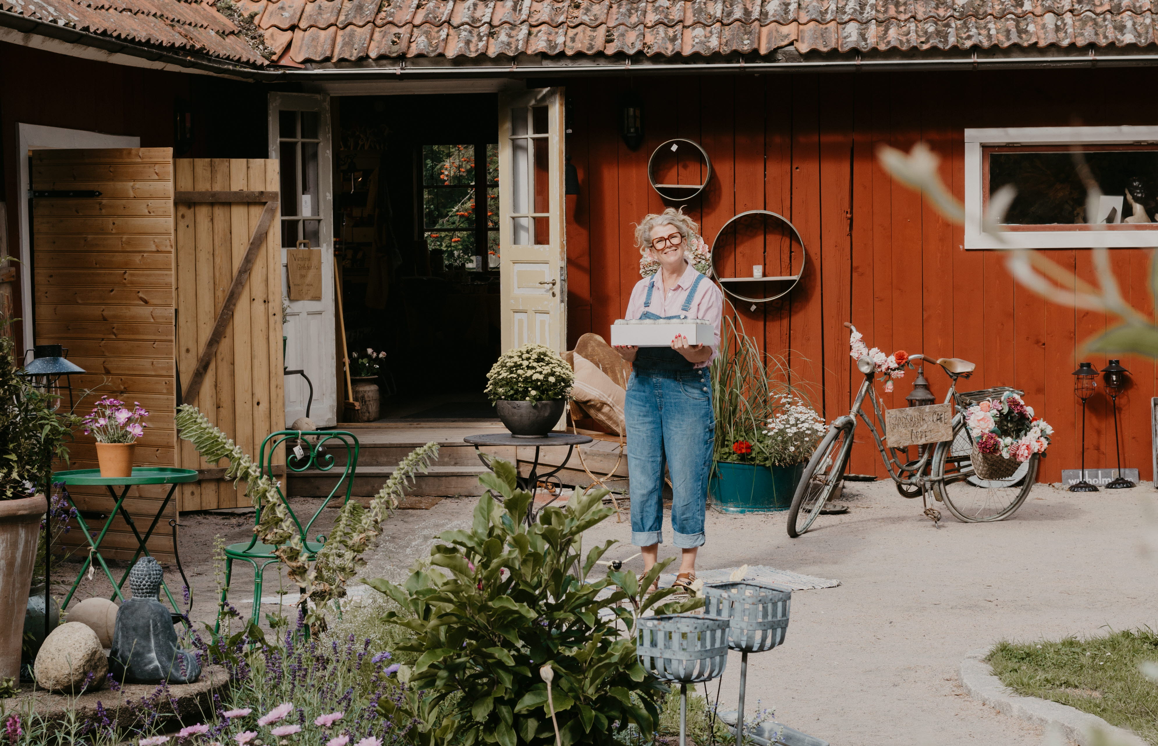 En kvinna håller i en låda. Hon har blå hängselbyxor på sig och står på en innergård. I förgrunden ser man en fin plantering.