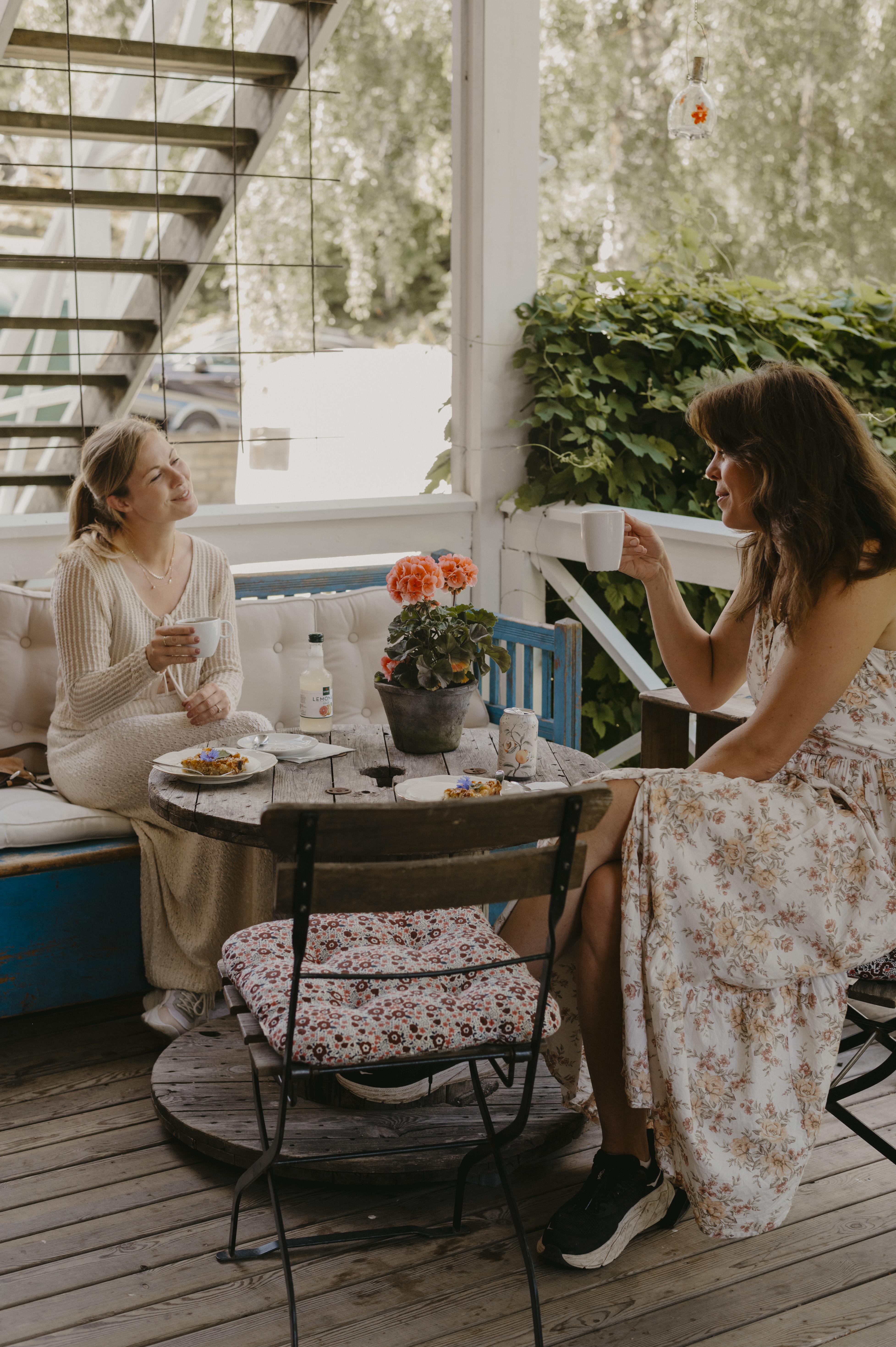 Två kvinnor i ljusa somriga kläder sitter på en altan och fikar.