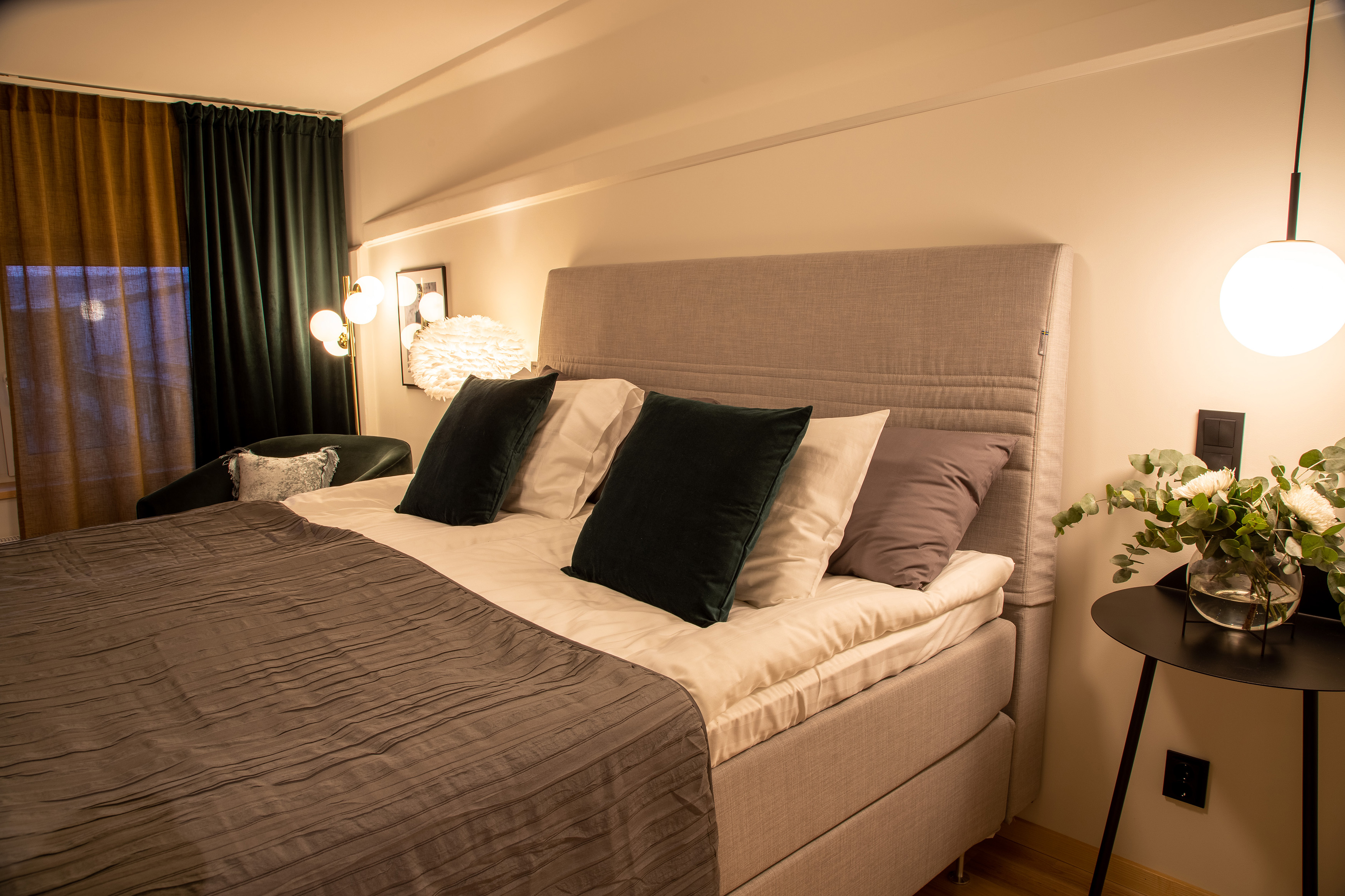 En säng med grå sänggavel i tyg. på ett bord bredvid står en vas med blommor.