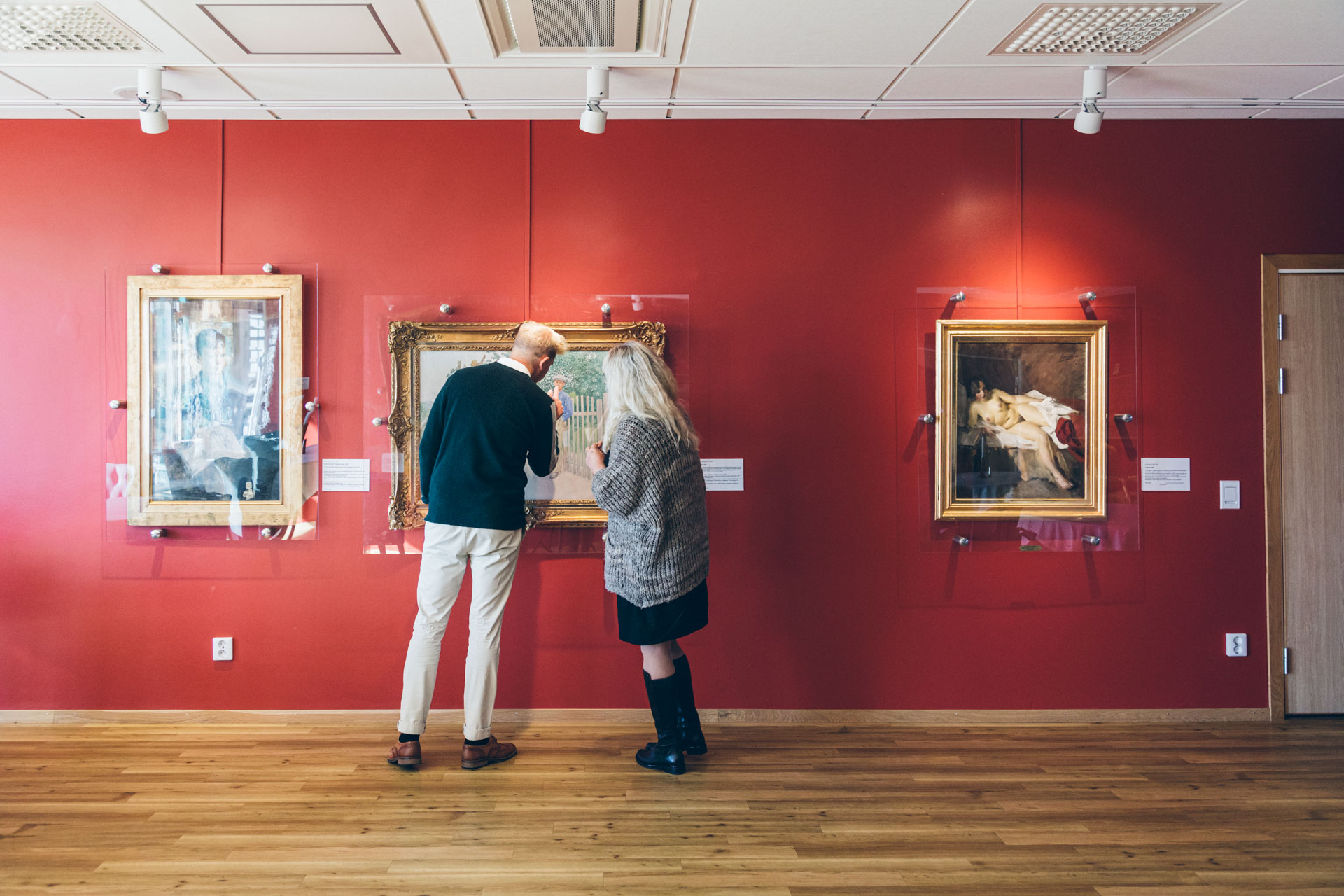 En kvinna i ljust hår och en man med kort ljust hår står i ett rum med röda väggar och tittar på konst. 