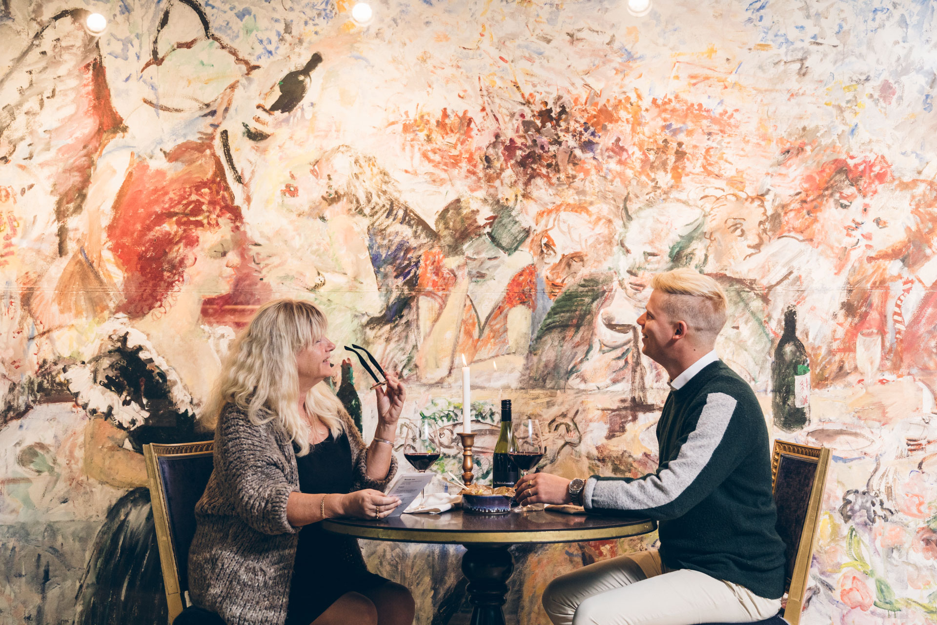 En kvinna i ljust hår och en man med kort ljust hår sitter vid ett bord med ett stort konstverk på väggen bredvid sig. 