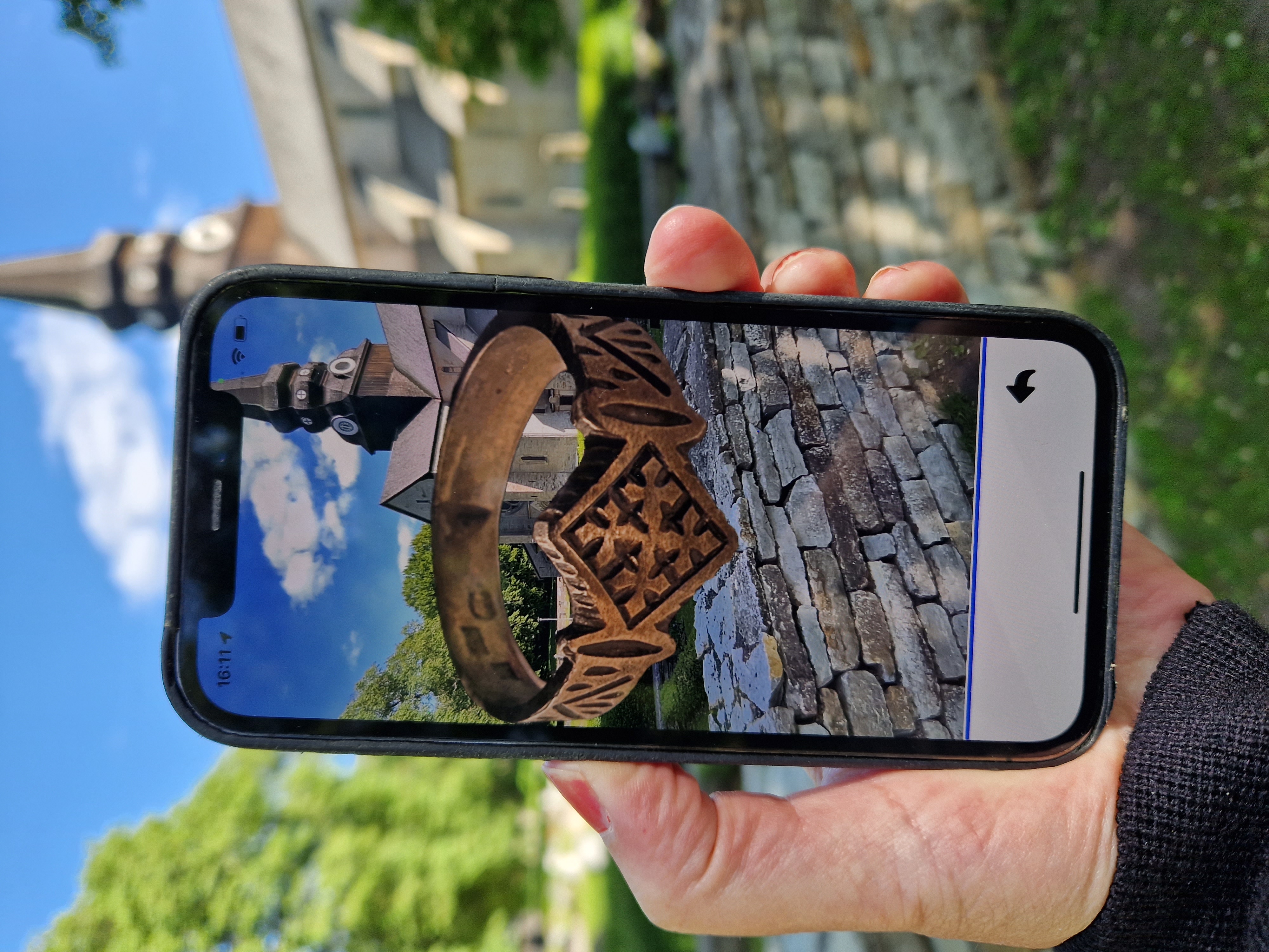 En hand som håller i en mobiltelefon. I bakgrunden syns en gammal kyrka och på mobilens skärm ser man en 3D-skannad ring.