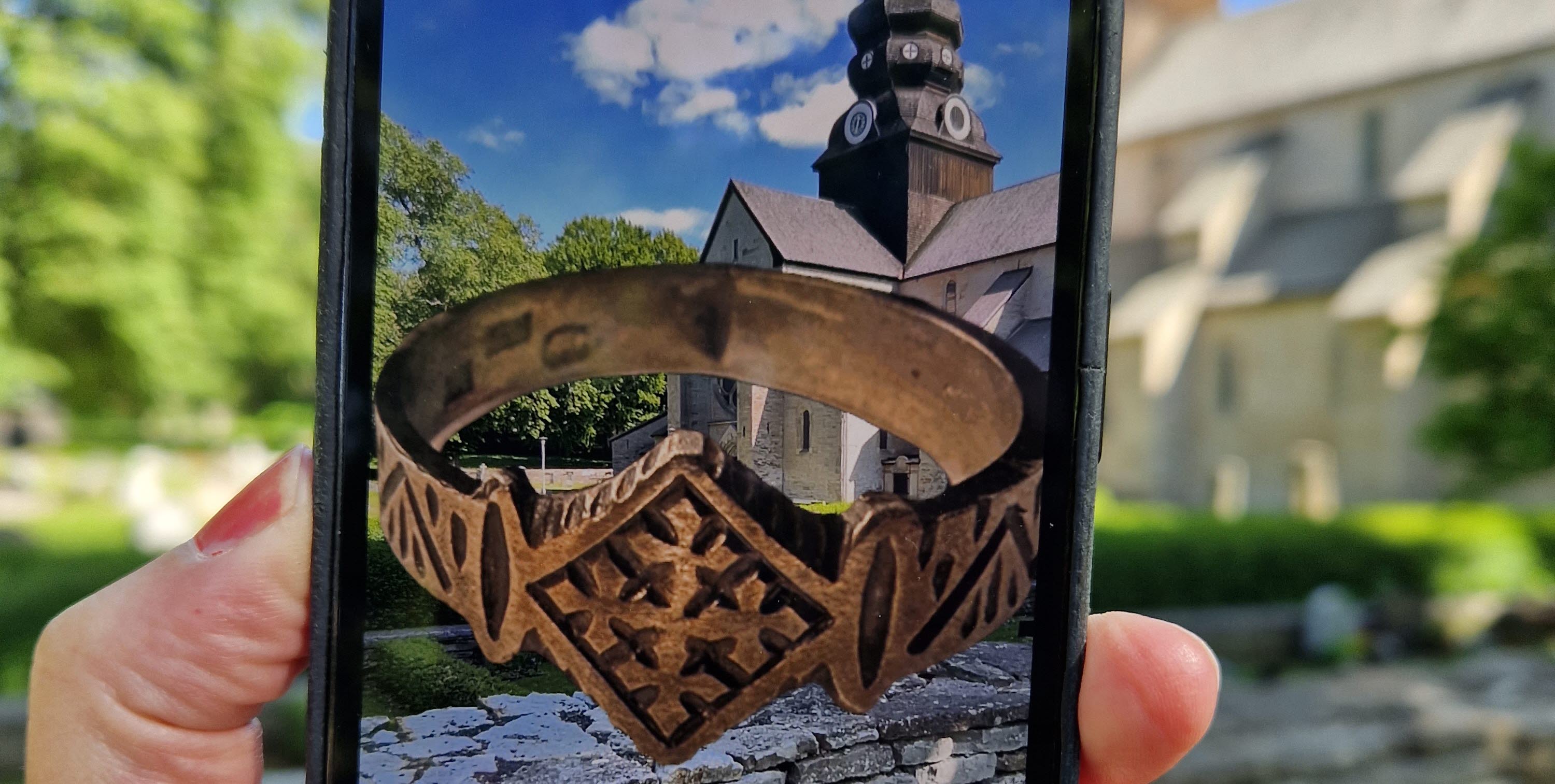 En hand som håller i en mobiltelefon. I bakgrunden syns en gammal kyrka och på mobilens skärm ser man en 3D-skannad ring.
