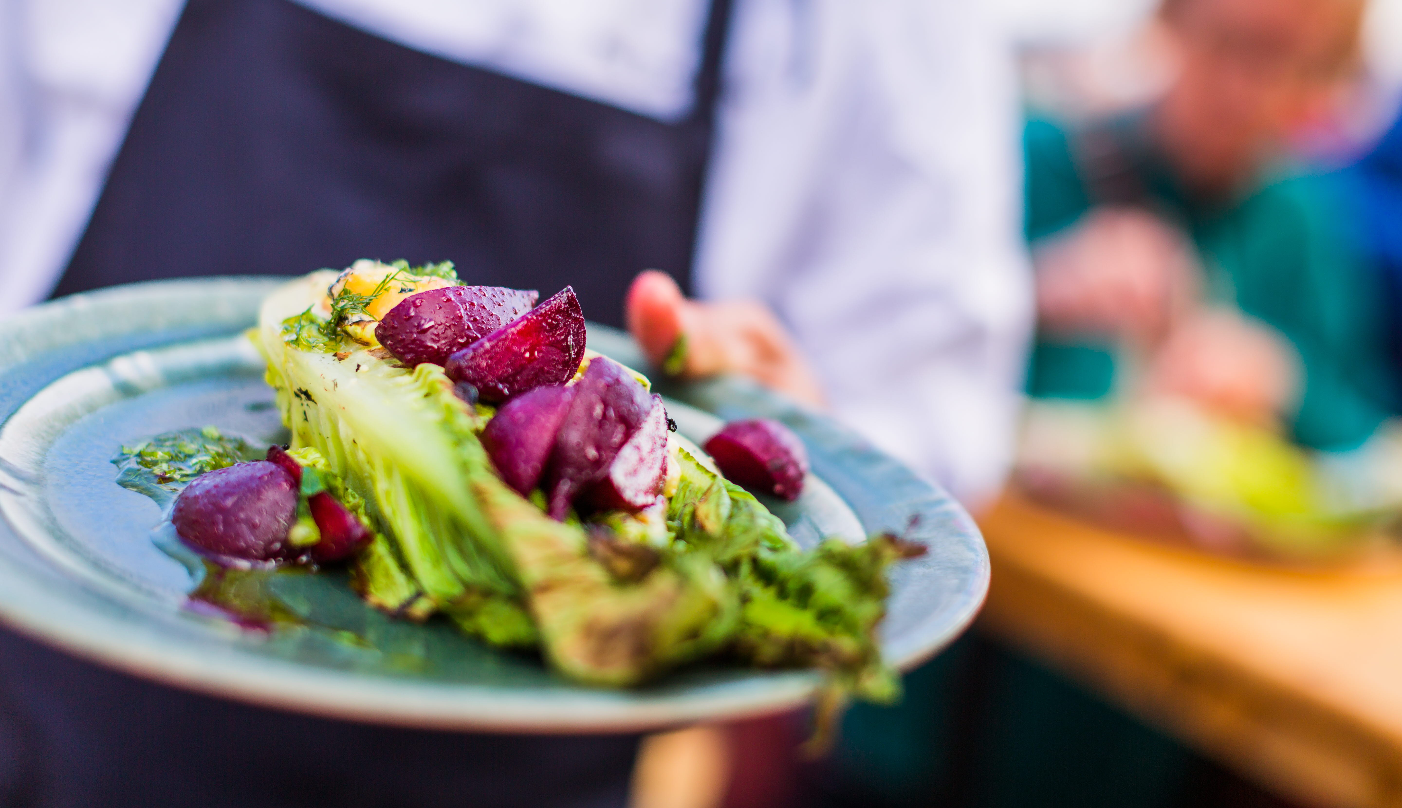 A person in white shirt and blue apron extends a turquoise plate with a colorful ingredients.