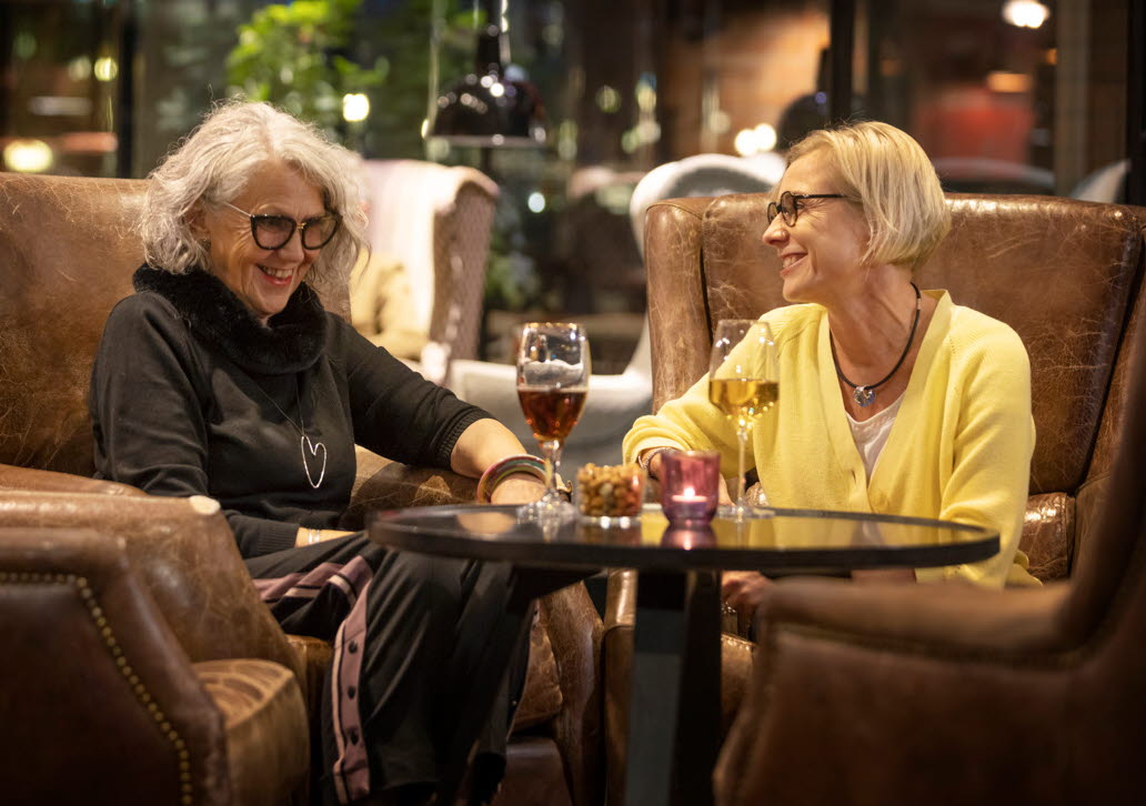 Två glada kvinnor i bruna fåtöljer på restaurang med varsitt glas vin. 