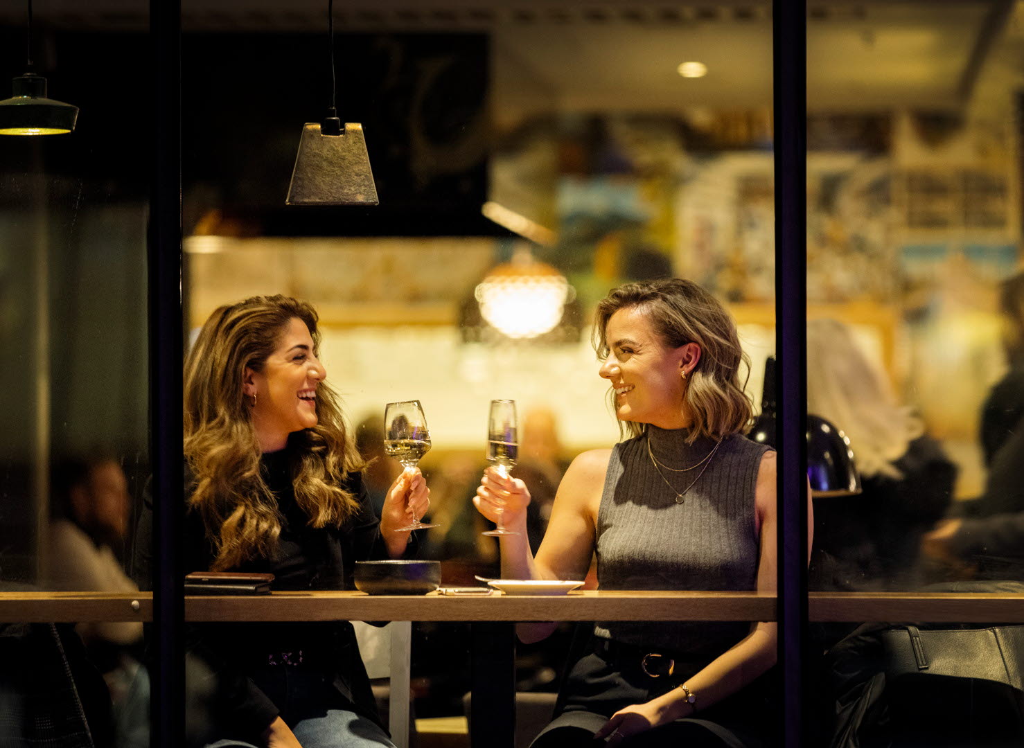 Två kvinnor sitter vid ett bord placerat vid ett fönster. De höjer varsitt glas mot varandra för att skåla. 