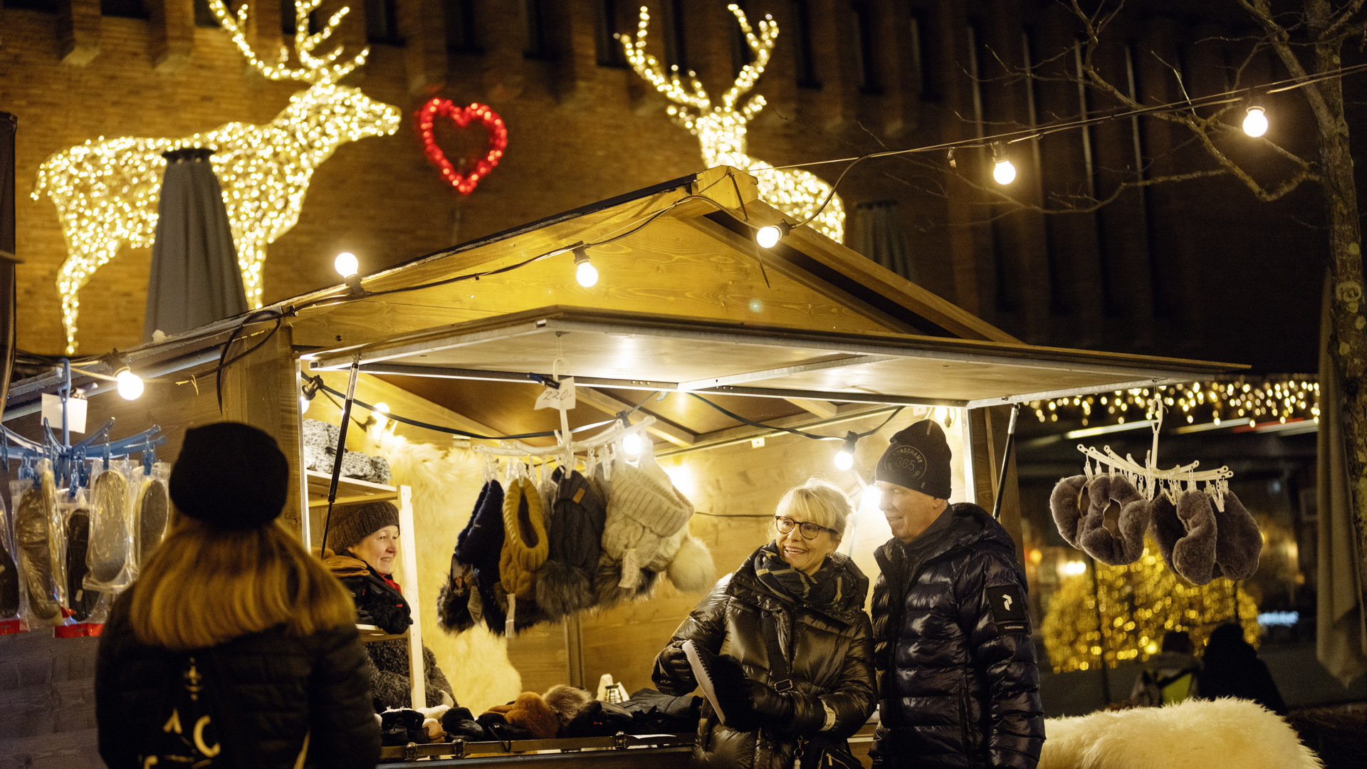 En äldre kvinna och äldre man klädda i svarta dunjackor står vid ett vackert upplyst marknadsstånd som säljer stickade mössor och vantar.  