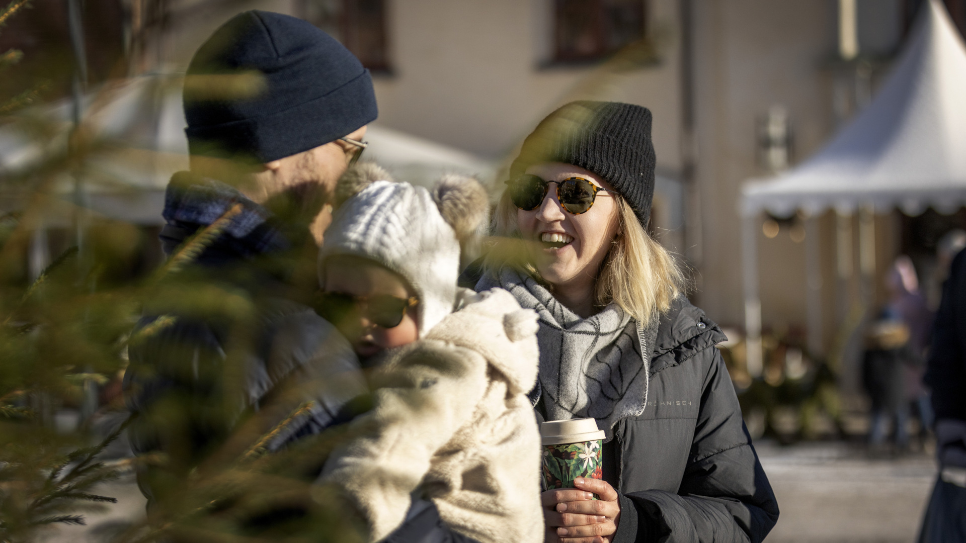 Ett yngre par står på torget i mörka mössor, svarta jackor och solglasögon. Mannen bär på ett barn i vit mössa med tofsar. Barnet har också solglasögonen. 