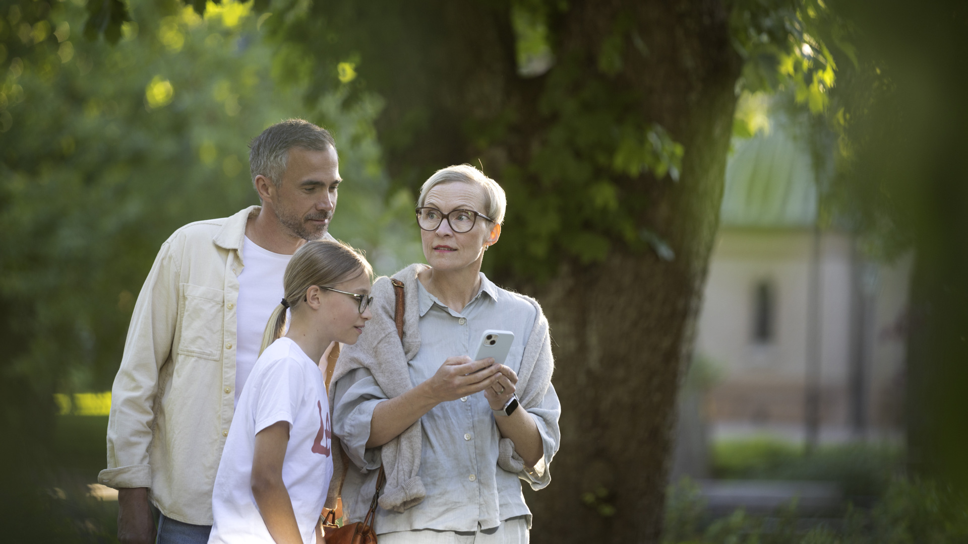 En man, en kvinna och en flicka står i en park och tittar ned i en telefon.