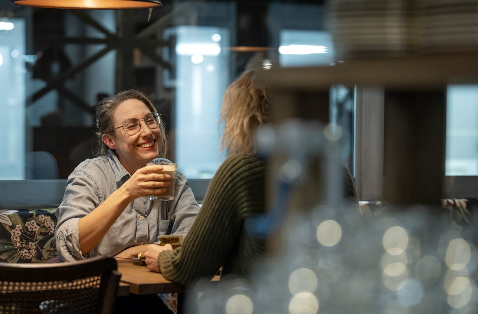 Två kvinnor sitter vid ett bord och fikar. En av dem ler och håller ett glas med kaffe i handen. 