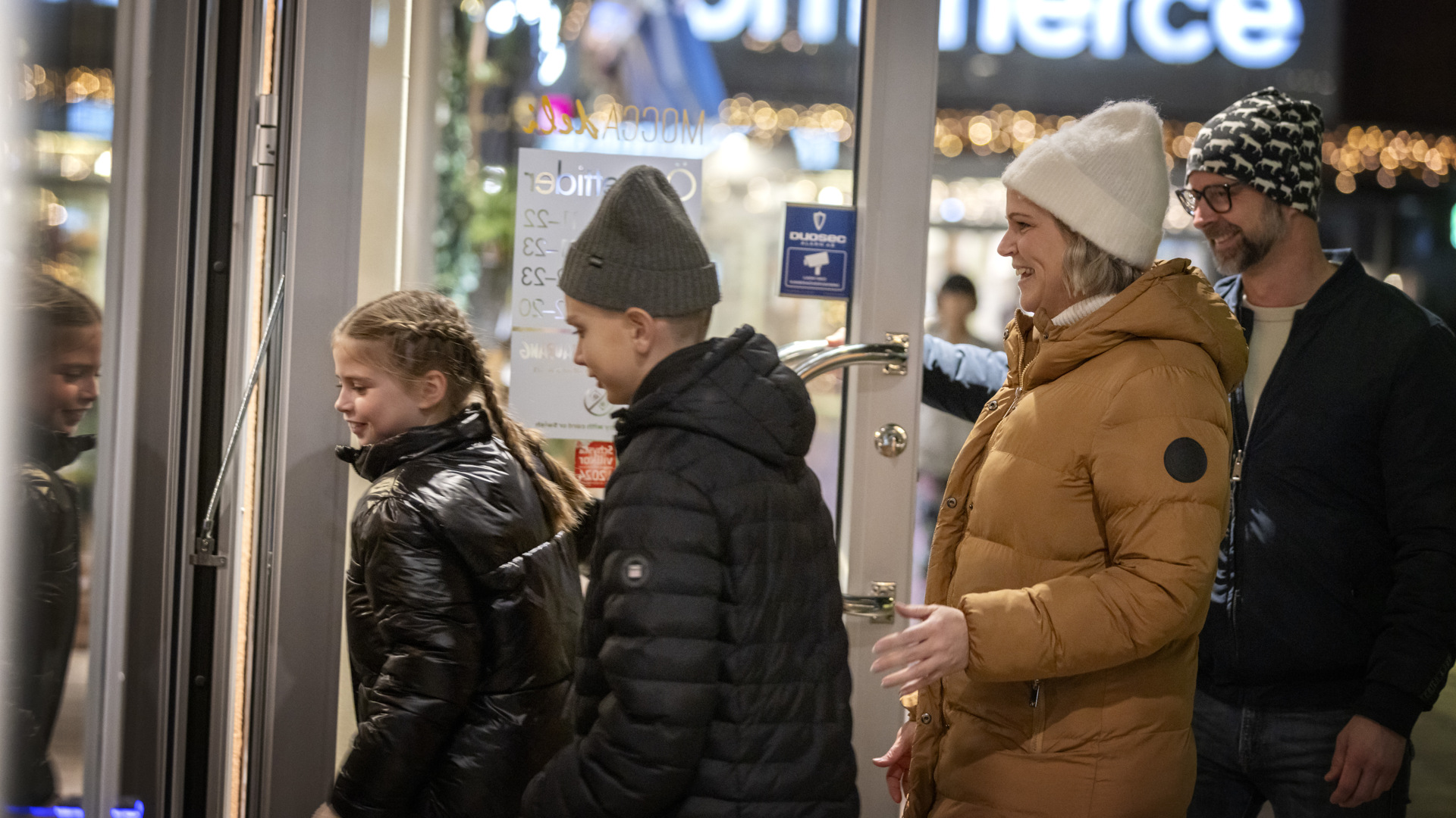 En kvinna i gul jacka är påväg in på en restaurang i sällskap med en man och två barn i svarta jackor. De ser alla glada ut. 