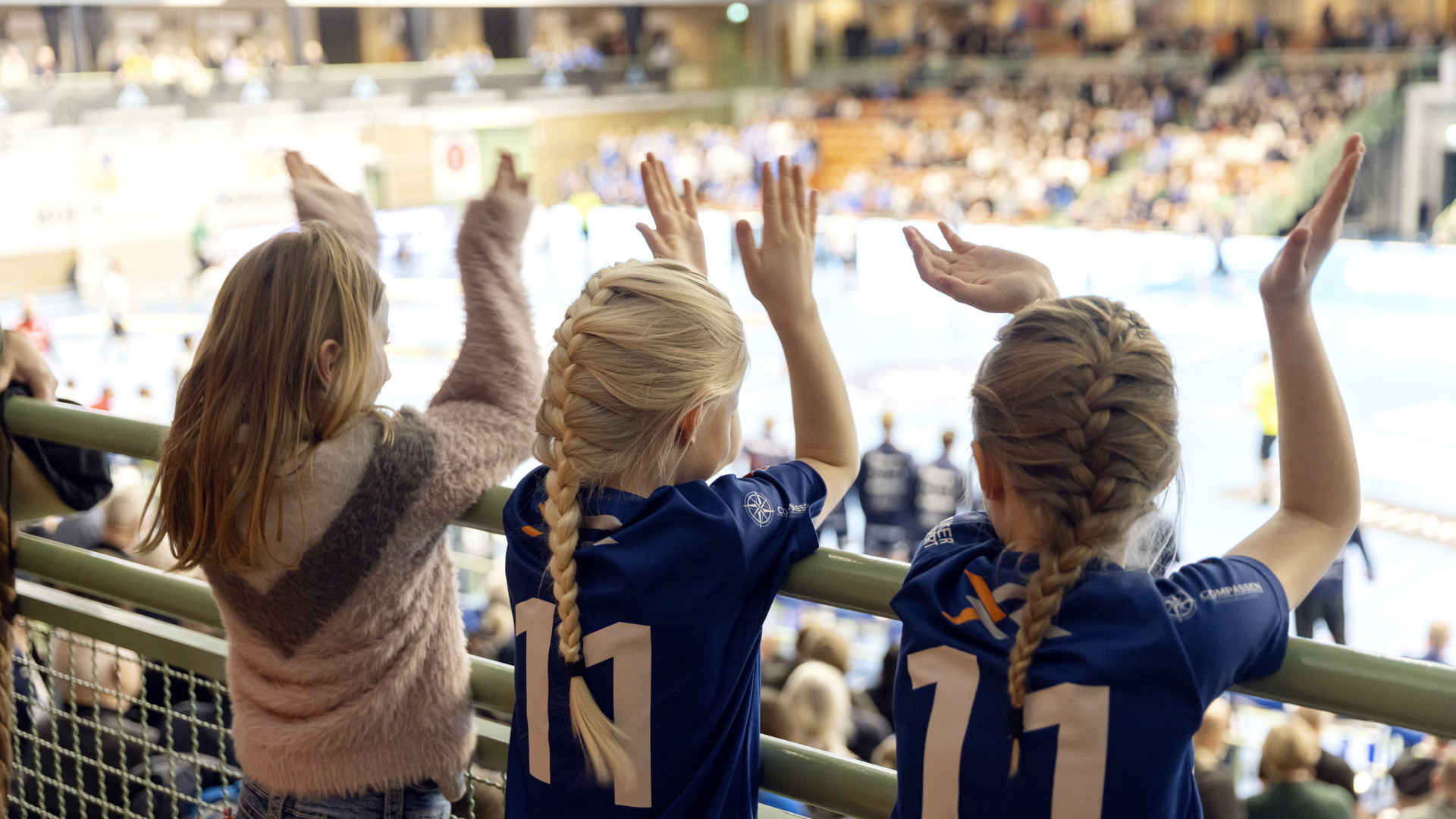 Tre tjejer står på rad på en läktare. De hejar och klappar med händerna medan de följer en pågående handbollsmatch. En tjej har håret utsläppt och de andra två har håret i fläta och bär varsin matchtröja. 