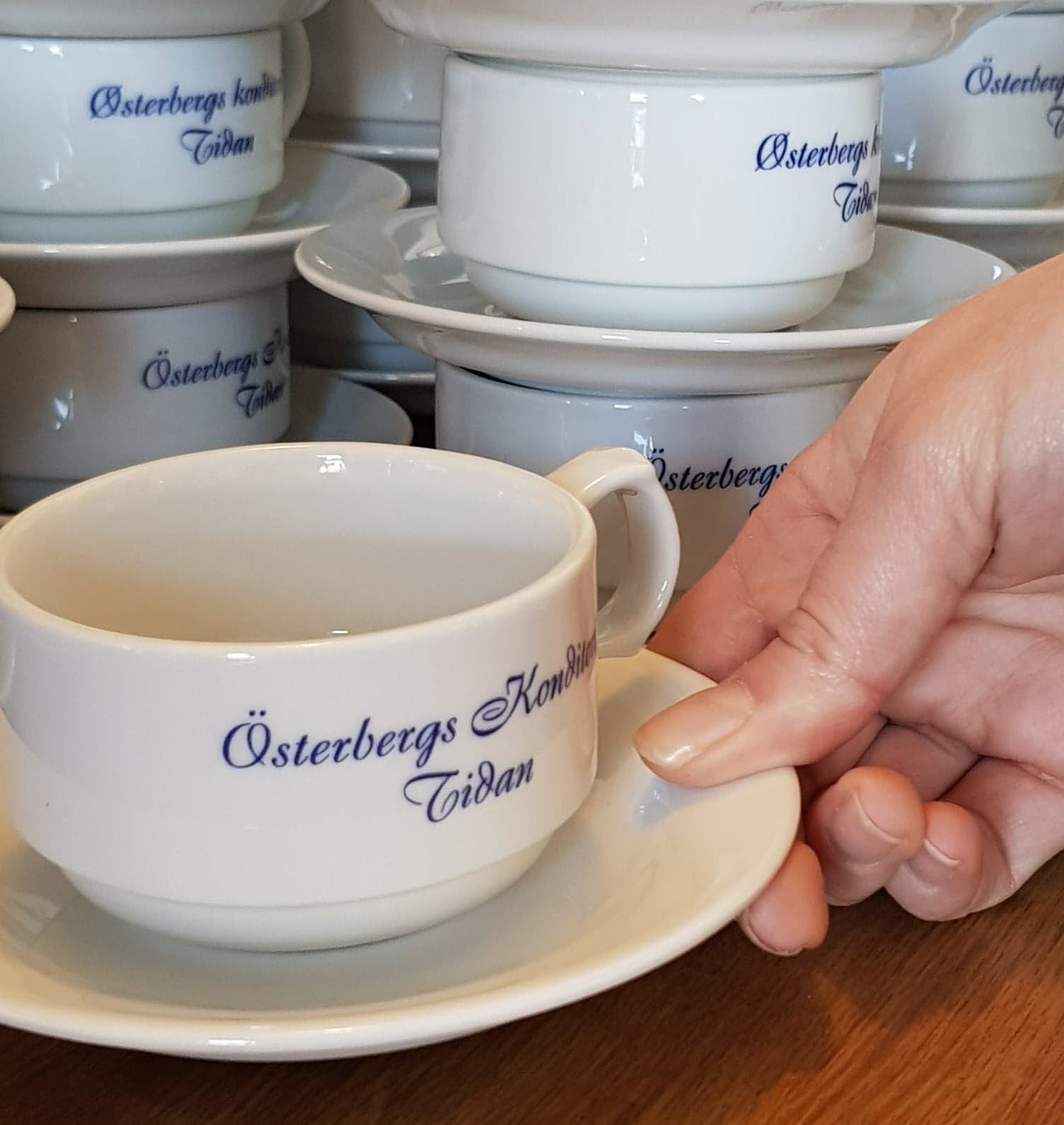 En hand lyfter en vit kopp med den snirkliga blå texten Österbergs Konditori Tidan
