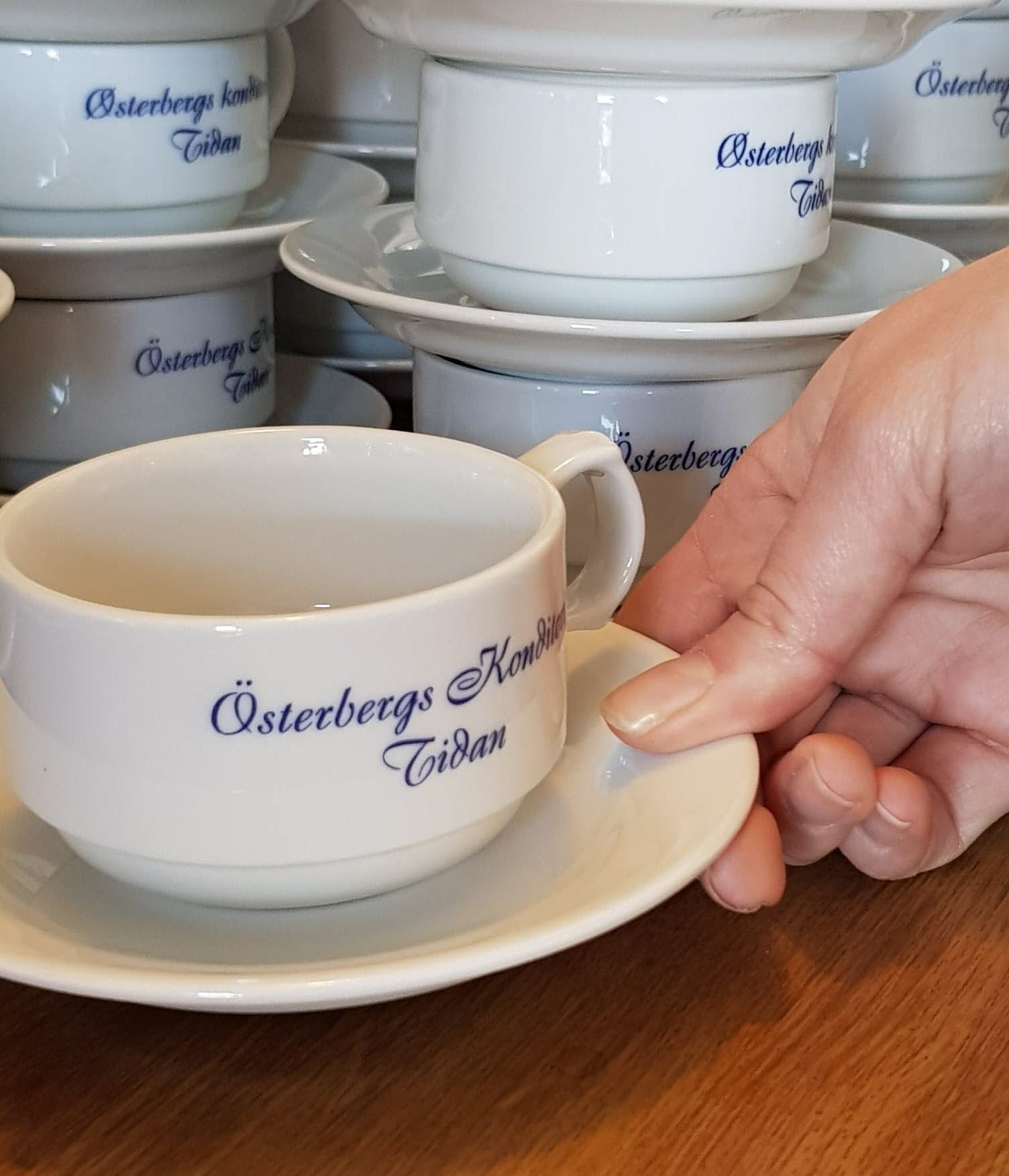 En hand lyfter en vit kopp med den snirkliga blå texten Österbergs Konditori Tidan