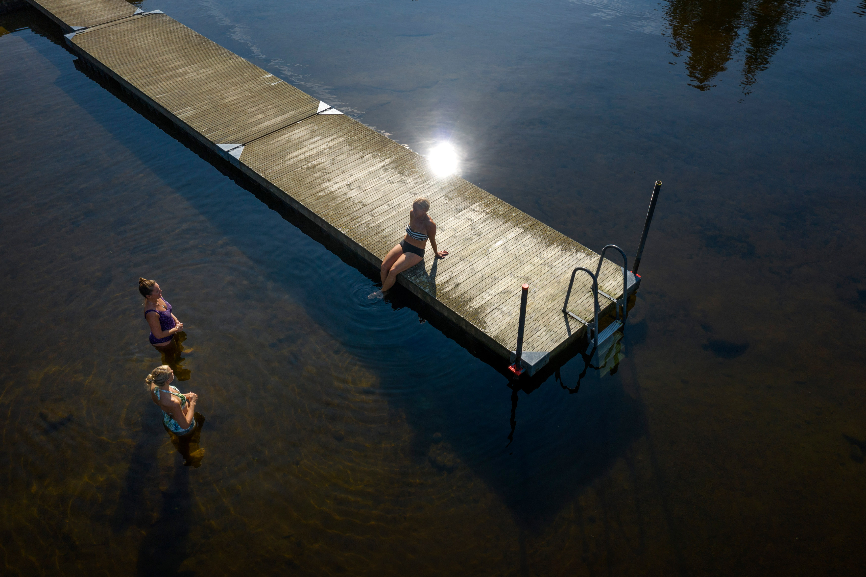Tre kvinnor badar i en sjö. En sitter på bryggan och pratar med de andra.